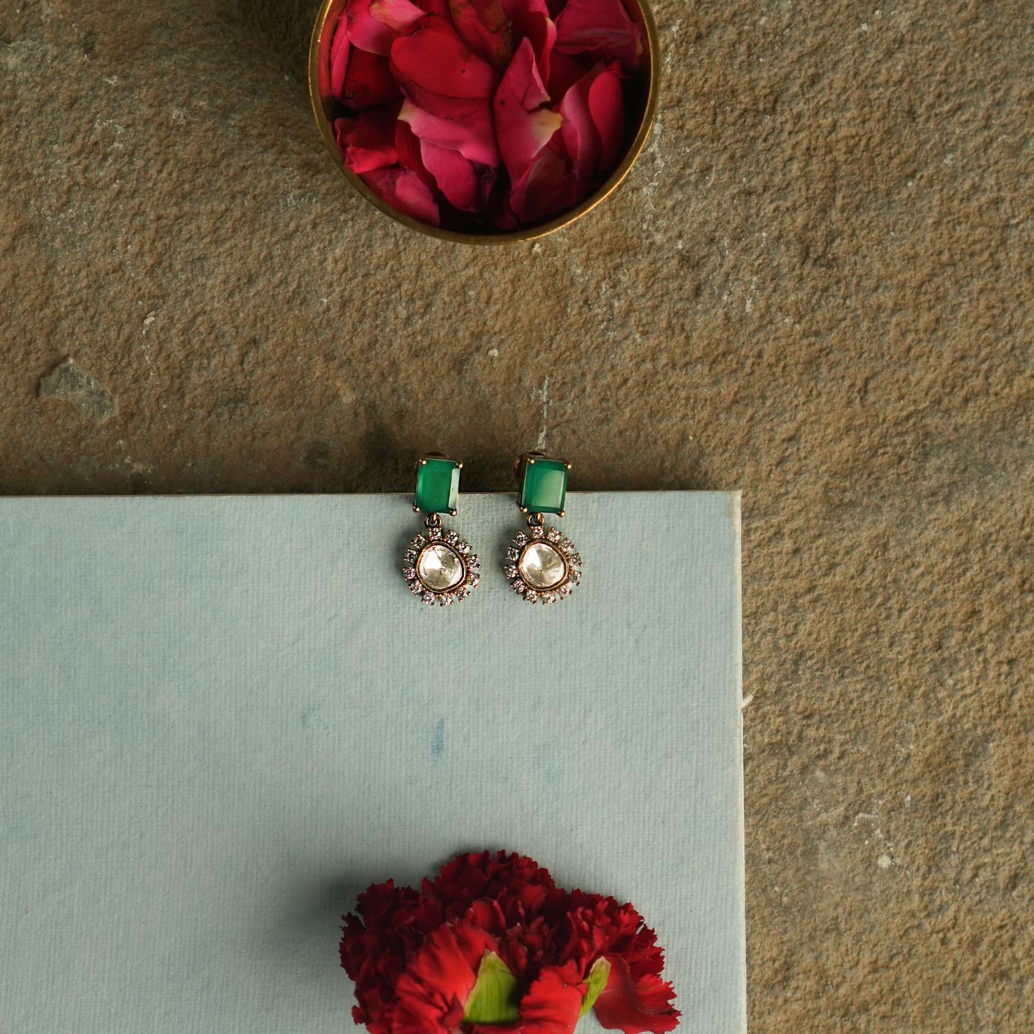 Amidst the vibrant charm of a bustling bazaar, this minimal floral stud with a radiant green cut stone at its heart, encircled by shimmering polki stones, whispers timeless elegance.