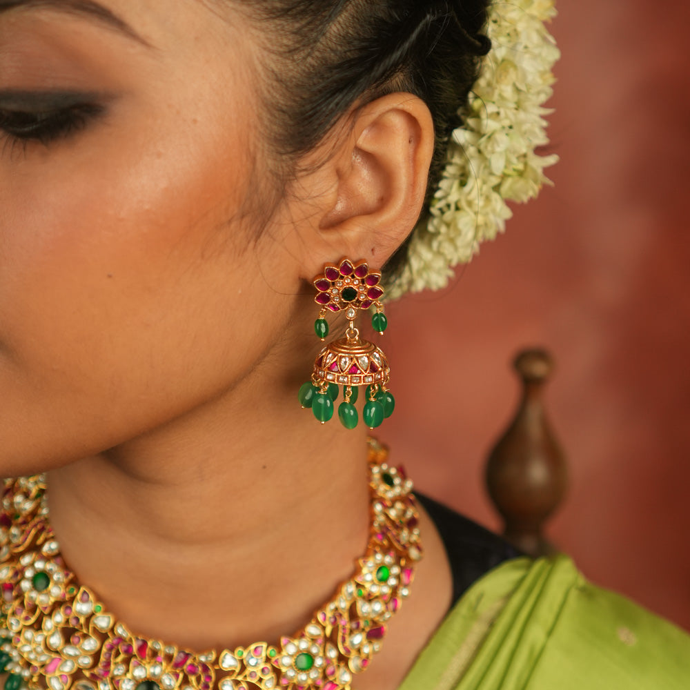 Regal charm reborn! These gold-plated jhumka earrings showcase radiant red and white Kundan stones, adorned with delicate green bead drops. A timeless symbol of celebration and exquisite artistry.