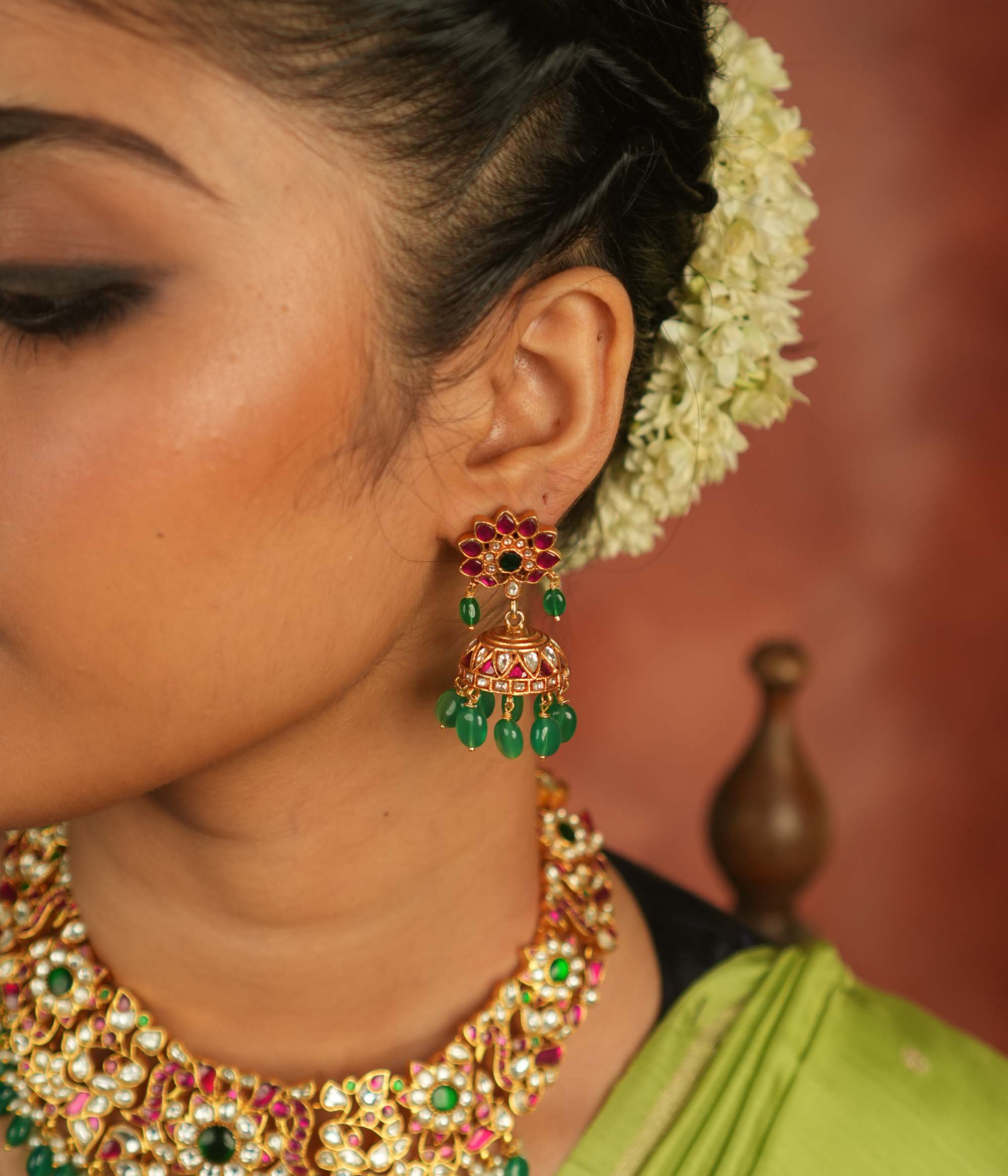 Regal charm reborn! These gold-plated jhumka earrings showcase radiant red and white Kundan stones, adorned with delicate green bead drops. A timeless symbol of celebration and exquisite artistry.