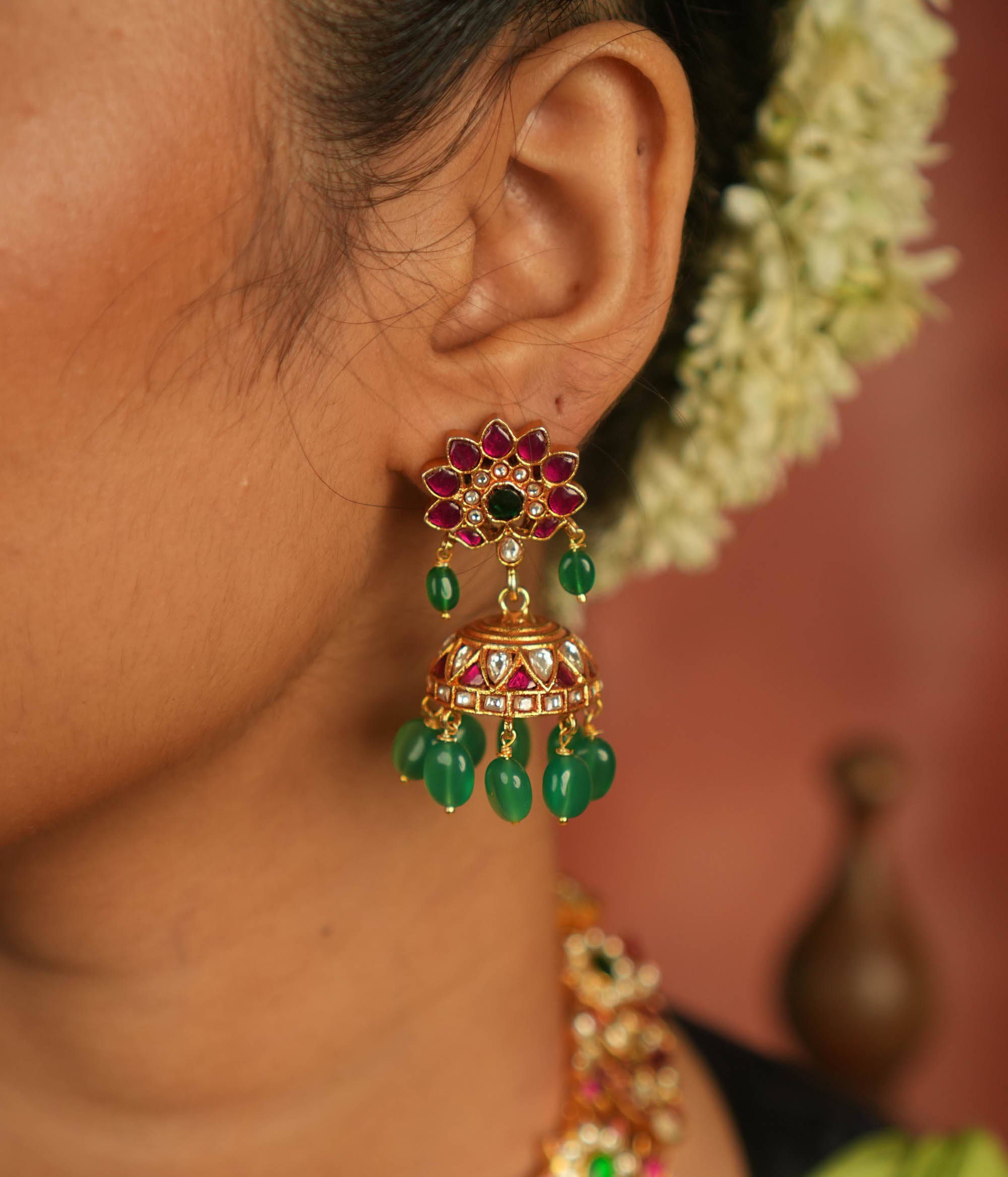 Regal charm reborn! These gold-plated jhumka earrings showcase radiant red and white Kundan stones, adorned with delicate green bead drops. A timeless symbol of celebration and exquisite artistry.