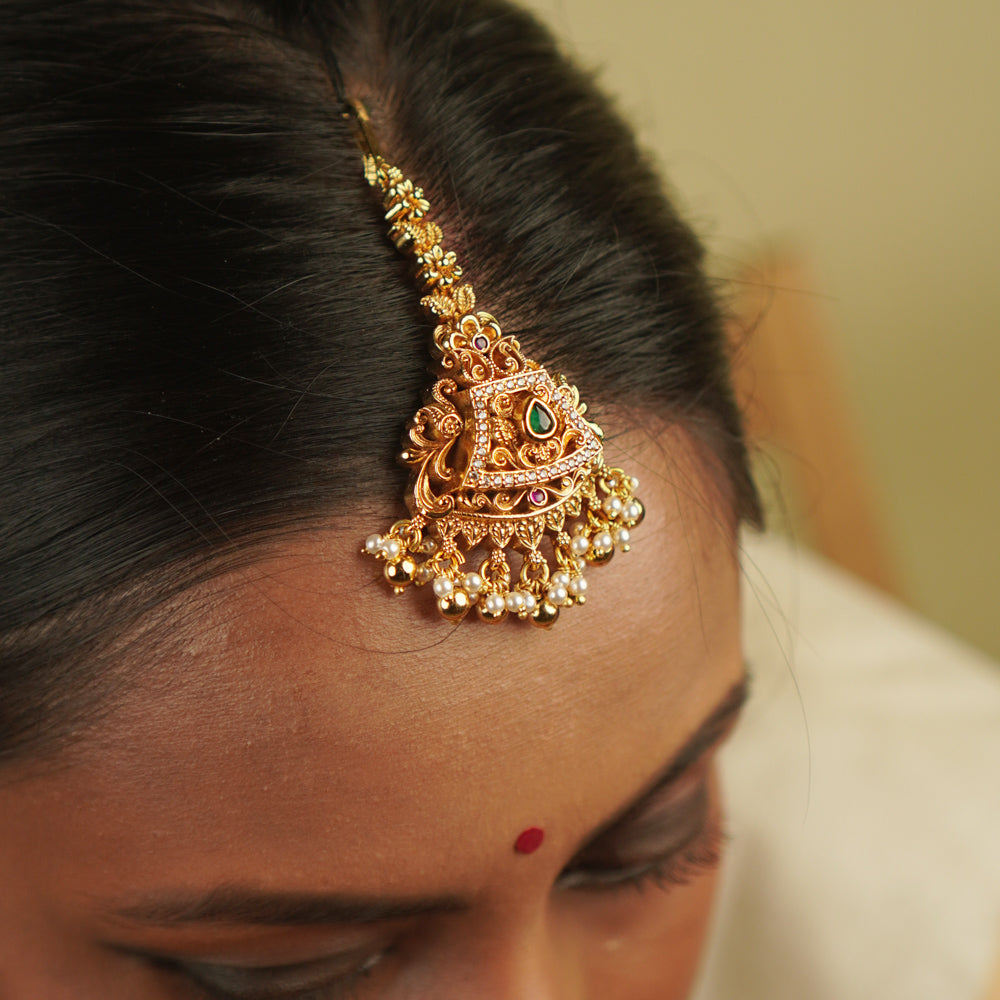 Yarima Maangtika: An embodiment of elegance. Crafted in gold-plated silver, this tikka features red and green spinel stones, CZ stones, and pearls, designed in a vintage Chettinad style. A cherished keepsake to affirm your timeless beauty.