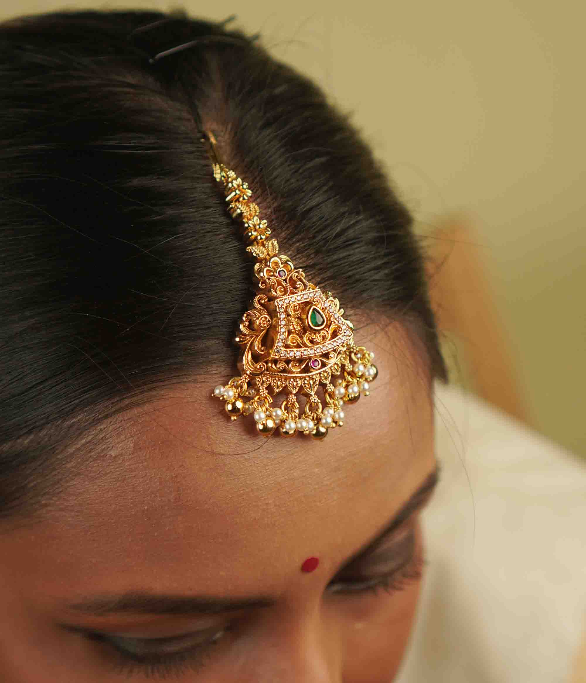 Yarima Maangtika: An embodiment of elegance. Crafted in gold-plated silver, this tikka features red and green spinel stones, CZ stones, and pearls, designed in a vintage Chettinad style. A cherished keepsake to affirm your timeless beauty.