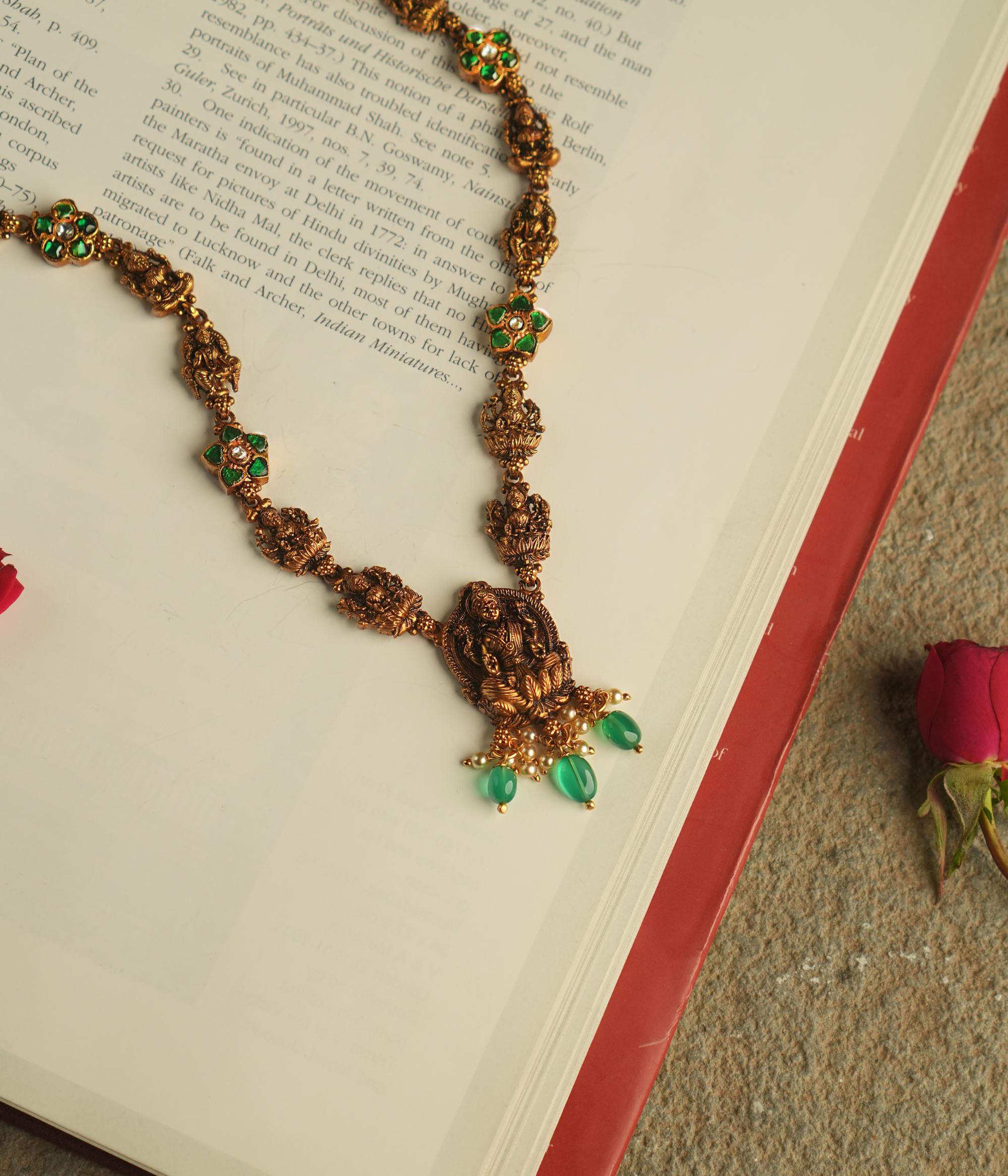 The Aadhya necklace evokes the charm of a bygone era, with antique gold, polkis, and a divine Lakshmi pendant. Pearl drops and green beads add a touch of timeless grace.