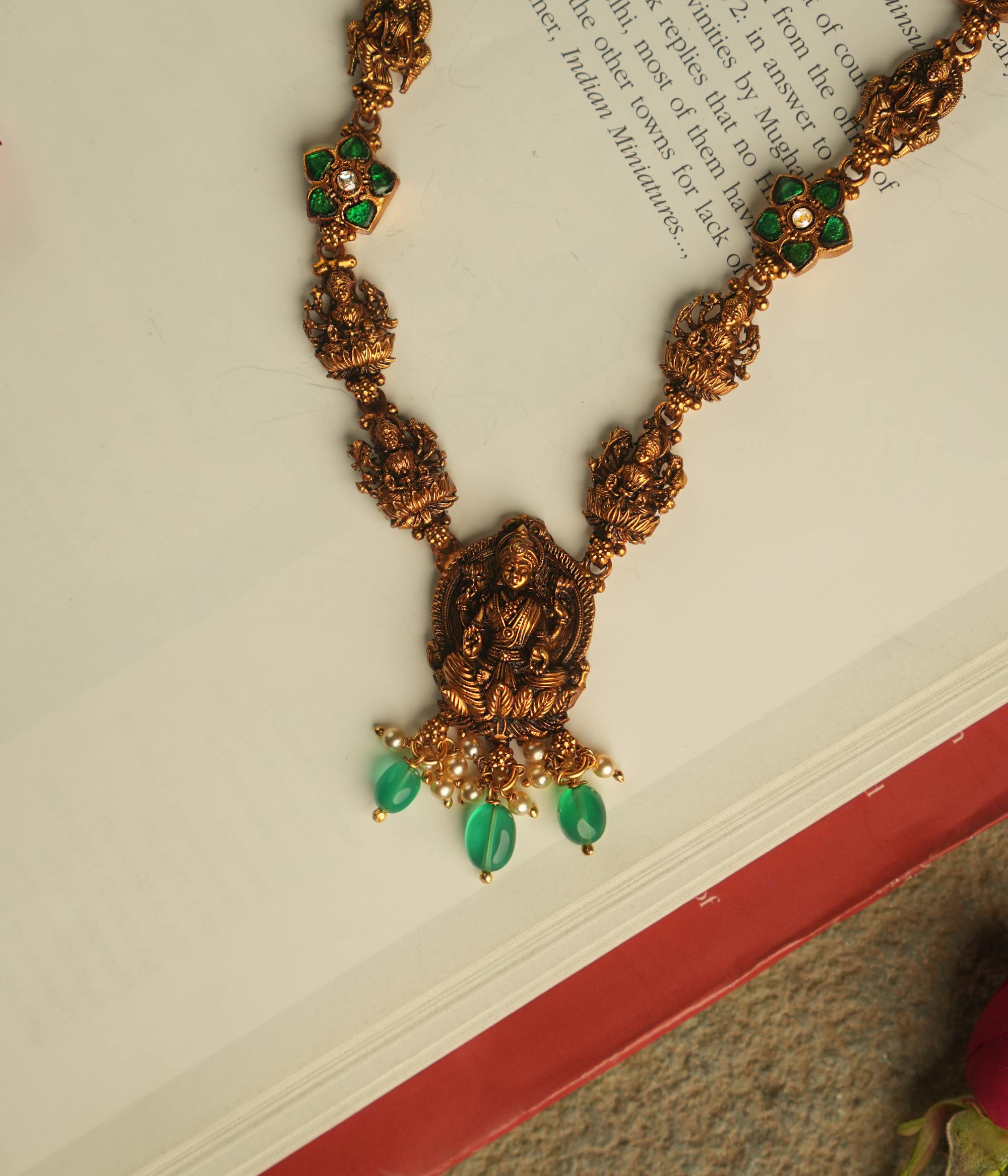 The Aadhya necklace evokes the charm of a bygone era, with antique gold, polkis, and a divine Lakshmi pendant. Pearl drops and green beads add a touch of timeless grace.
