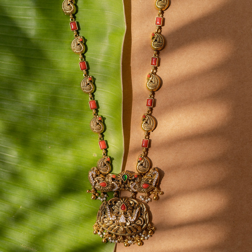 A 92.5 sterling silver Mythical Peacocks Antique Temple Necklace, likely gold-plated, featuring twin peacocks and a yali motif accented with red and green kempu stones, with a chain alternating between peacock links and coral-hued rectangular elements, a perfect heirloom-style piece for festive and bridal occasions.