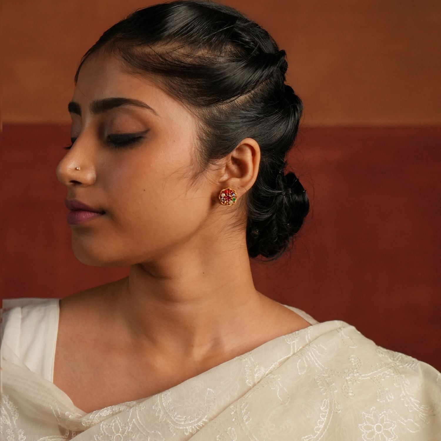 A beautifully crafted gold-plated silver stud earring in a delicate round shape, featuring vibrant Navratna stones arranged in a floral design. The center is adorned with a dazzling CZ stone, enhancing its elegance. This timeless piece blends tradition with sophistication, making it a perfect accessory for any occasion.
