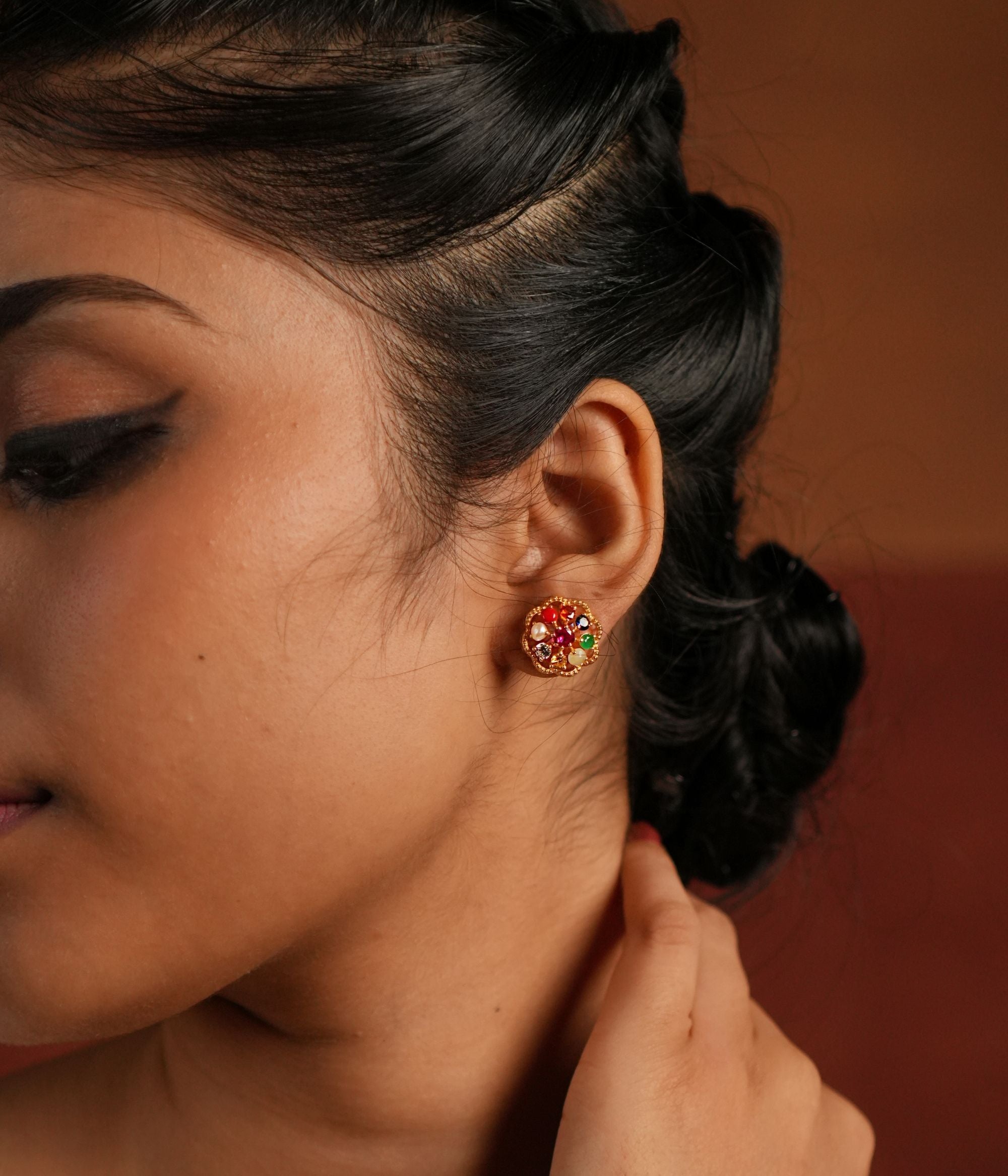 A beautifully crafted gold-plated silver stud earring in a delicate round shape, featuring vibrant Navratna stones arranged in a floral design. The center is adorned with a dazzling CZ stone, enhancing its elegance. This timeless piece blends tradition with sophistication, making it a perfect accessory for any occasion.