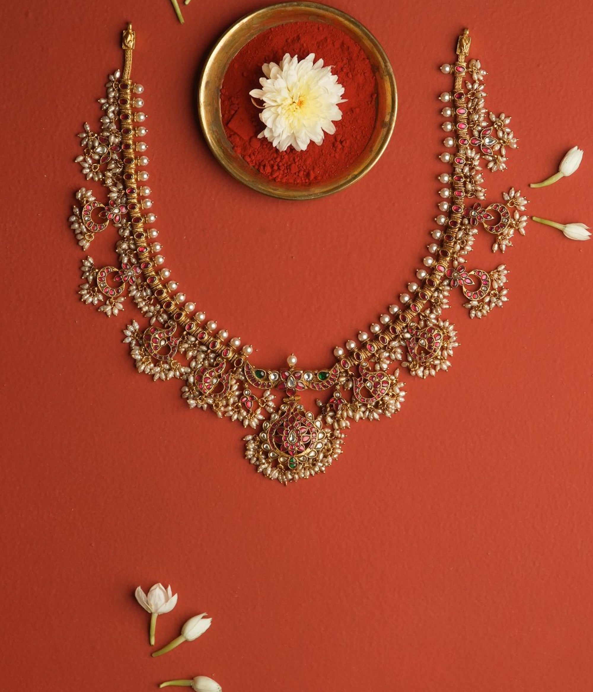 Ornate gold necklace on a red background with a small bowl and flowers.