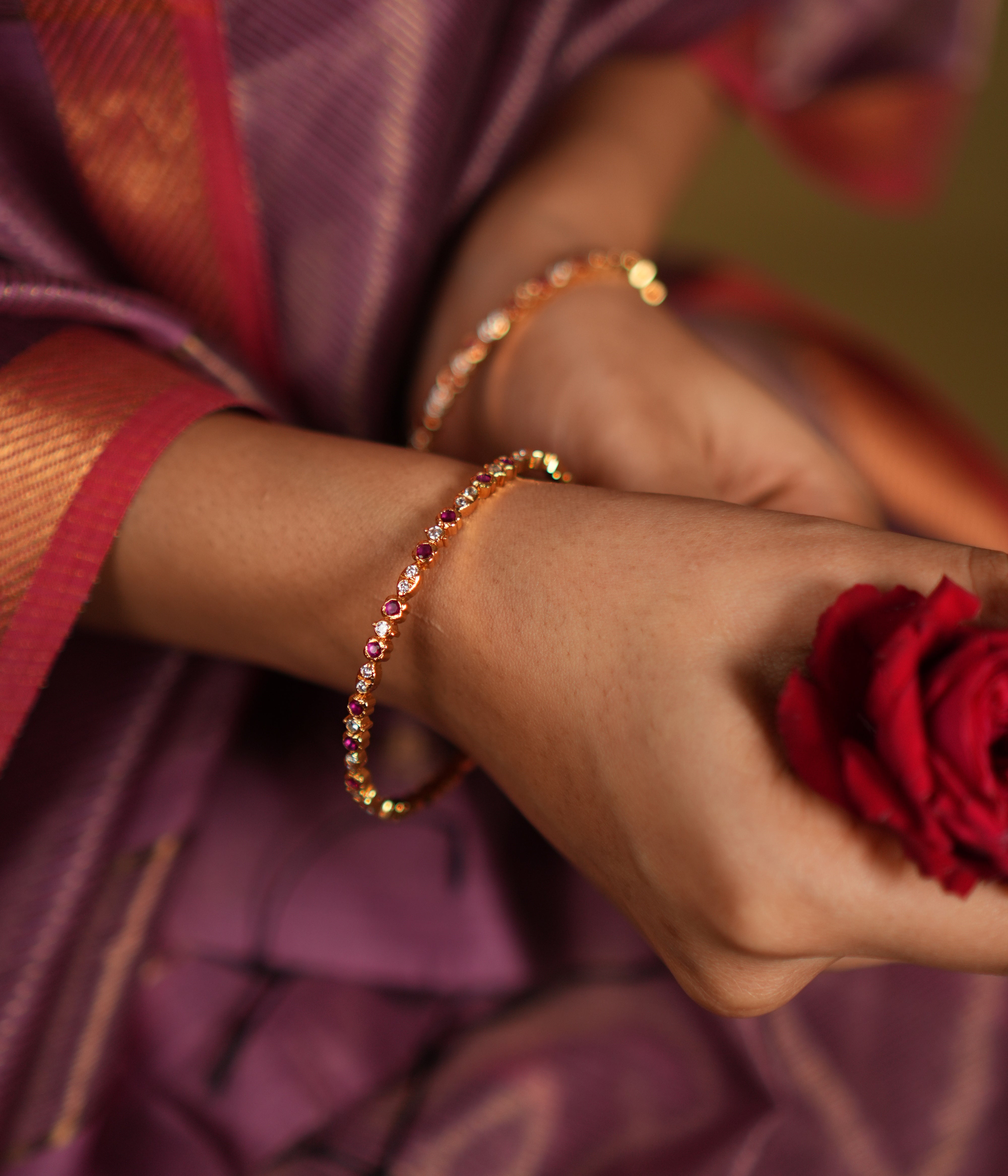 Close-up of a woman's wrist adorned with elegant silver bangles, gold-plated and embellished with white and red cubic zirconia stones, complemented by a vibrant red rose and a traditional silk saree in shades of purple and red. 