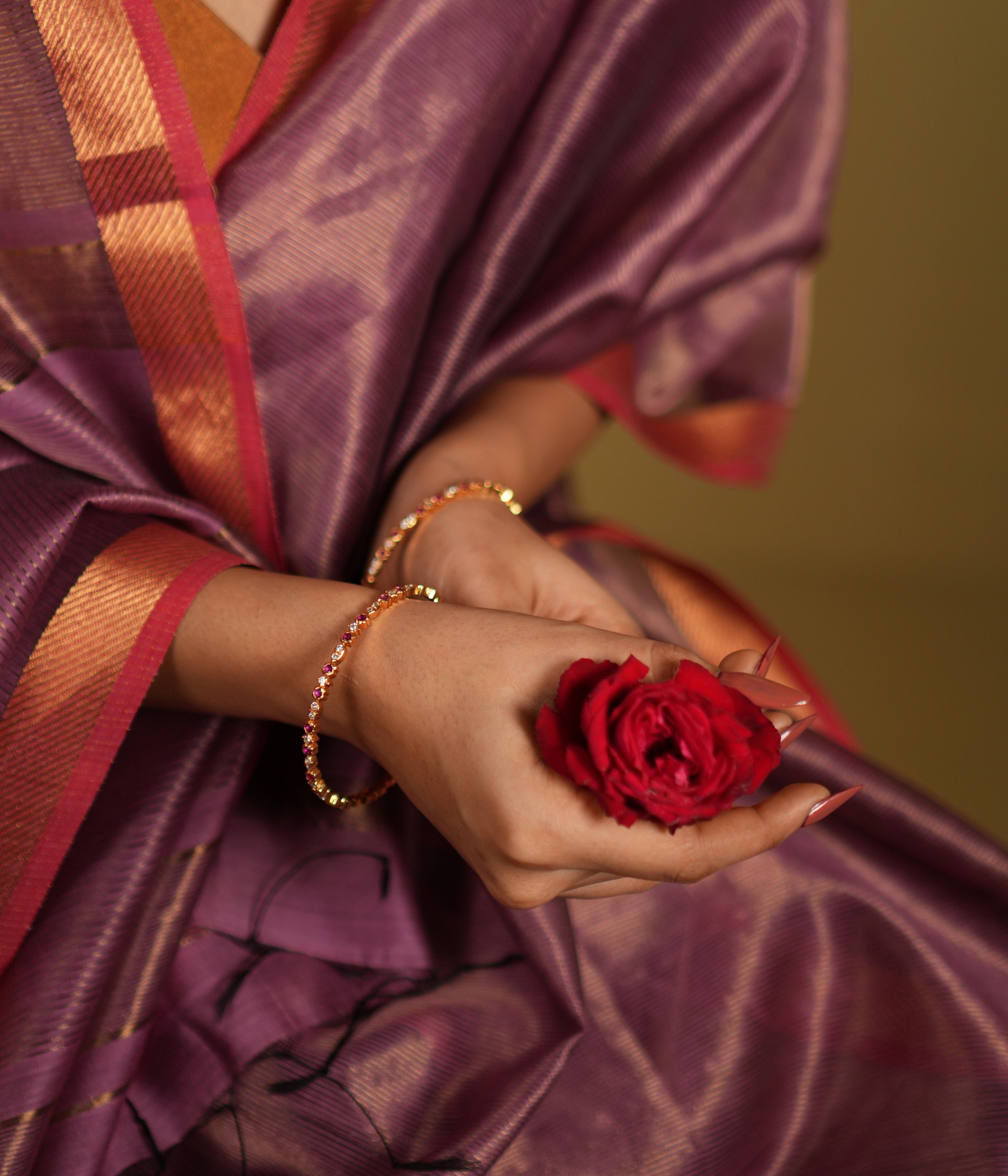 Close-up of a woman's wrist adorned with elegant silver bangles, gold-plated and embellished with white and red cubic zirconia stones, complemented by a vibrant red rose and a traditional silk saree in shades of purple and red. 