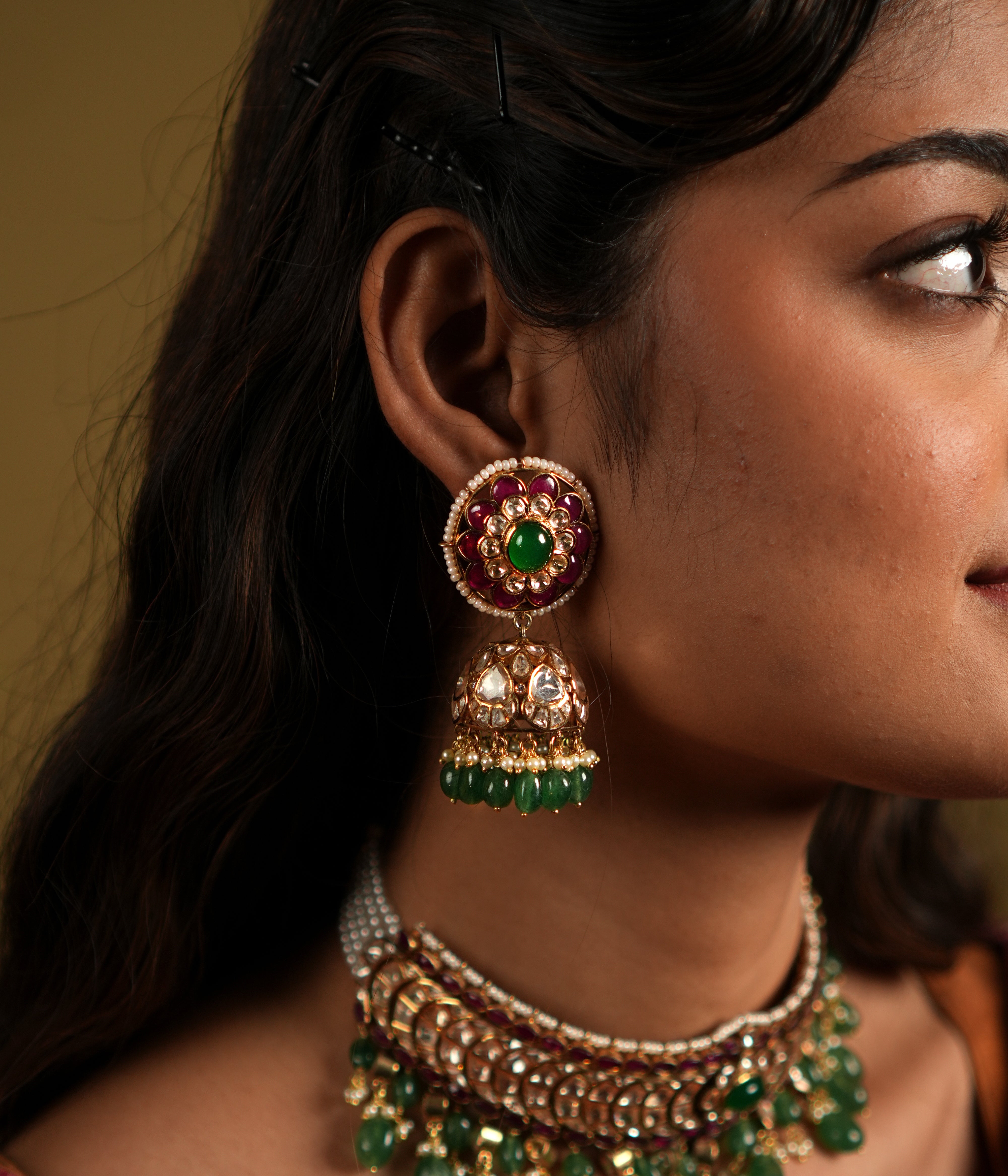 This image showcases an exquisite pair of Moissanite Polki Jhumkas crafted from 92.5 sterling silver and finished with a luxurious gold plating. The earrings feature a vibrant combination of deep red and green stones, with intricate Polki work that exudes traditional elegance.