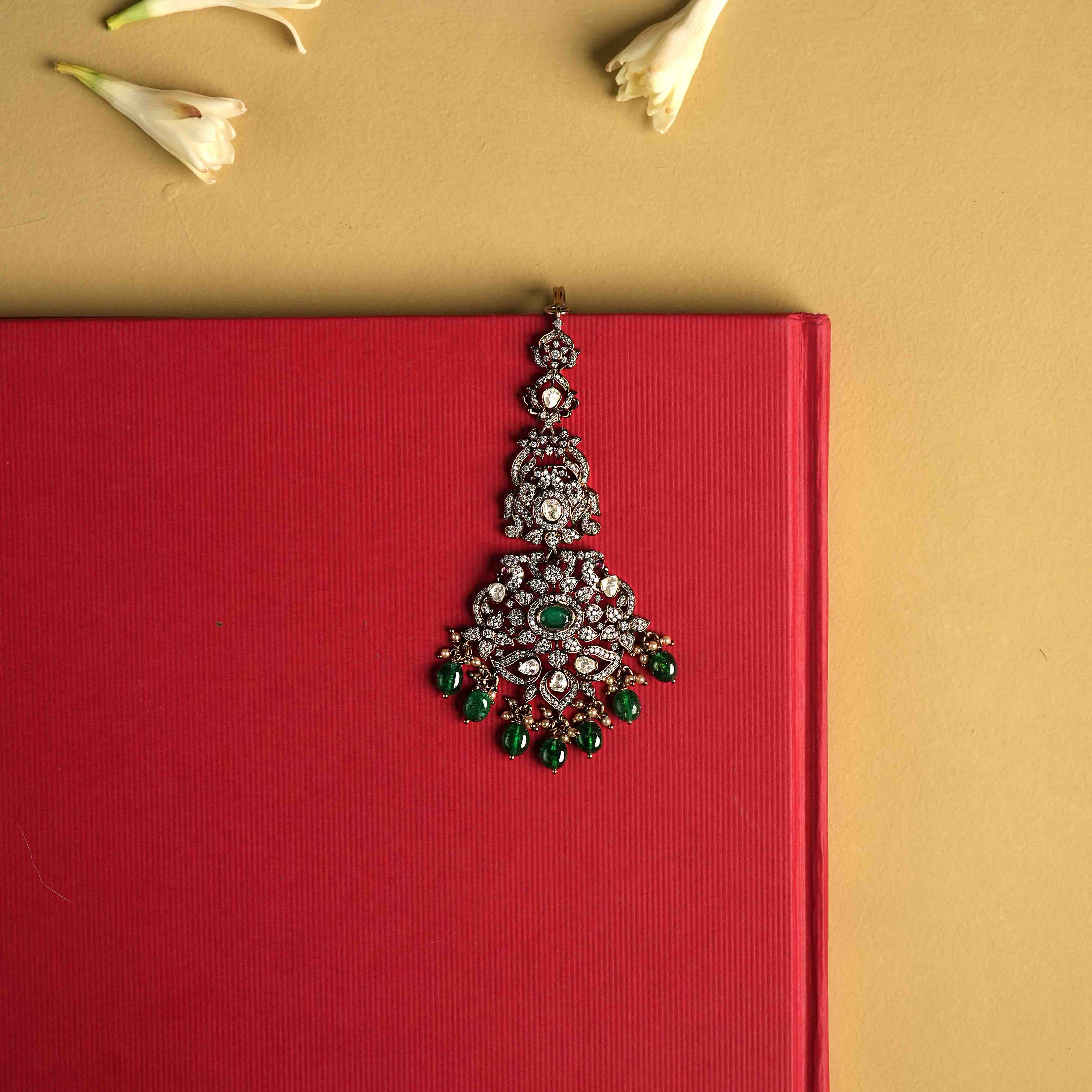 This intricately crafted Maangtika, handmade from 92.5 sterling silver with 14ct dark antique gold plating, features polki stones, CZ stones, green gemstones, green beads, and pearls.  The design shines with polki and moissanite brilliance, with cultured emerald dangles adding a touch of majestic elegance, evoking the opulence of a Victorian era.