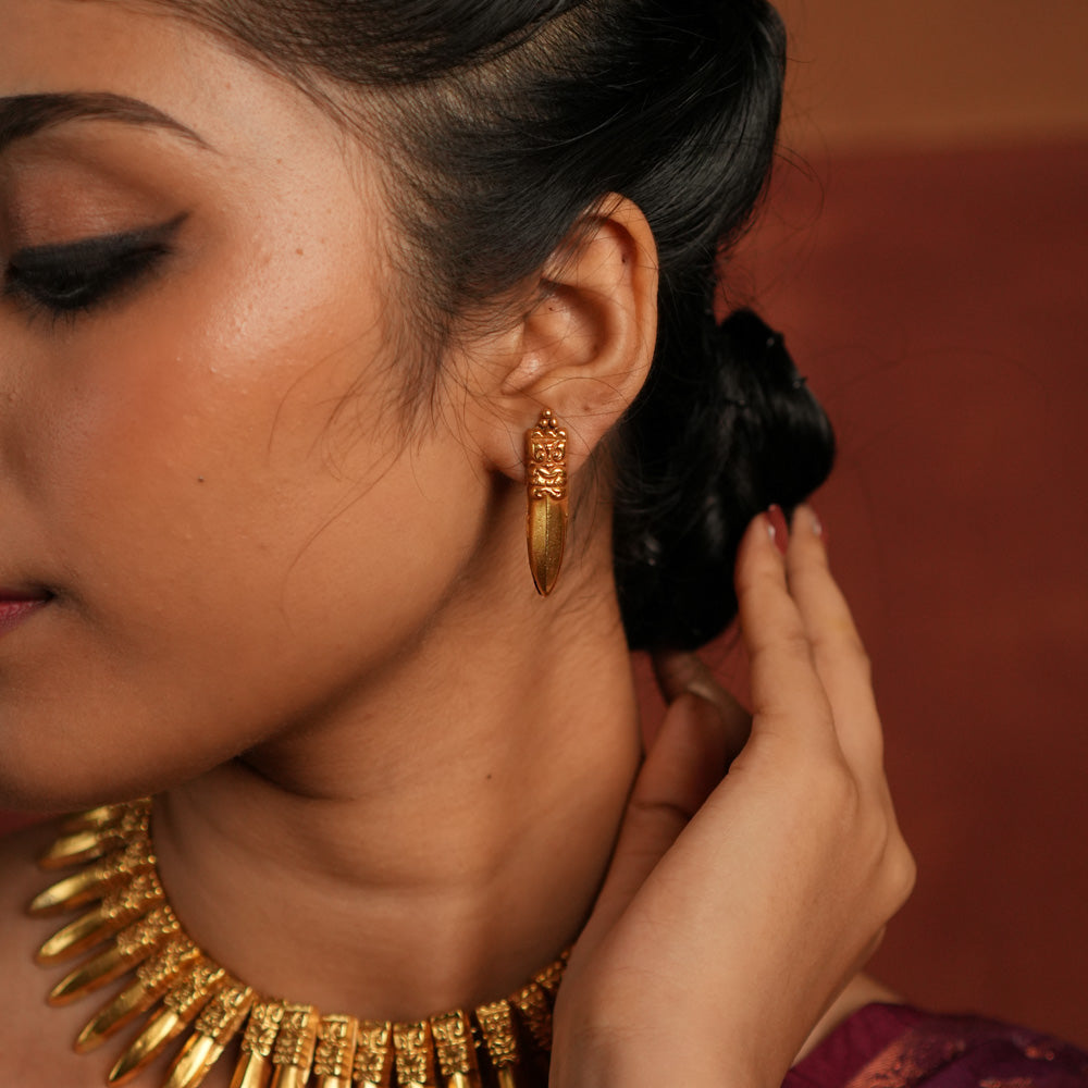 A close-up of a gold-plated earring inspired by traditional Indian designs, featuring an intricately carved top with a fierce face motif and a sleek, pointed pendant drop, worn with an elegant updo hairstyle