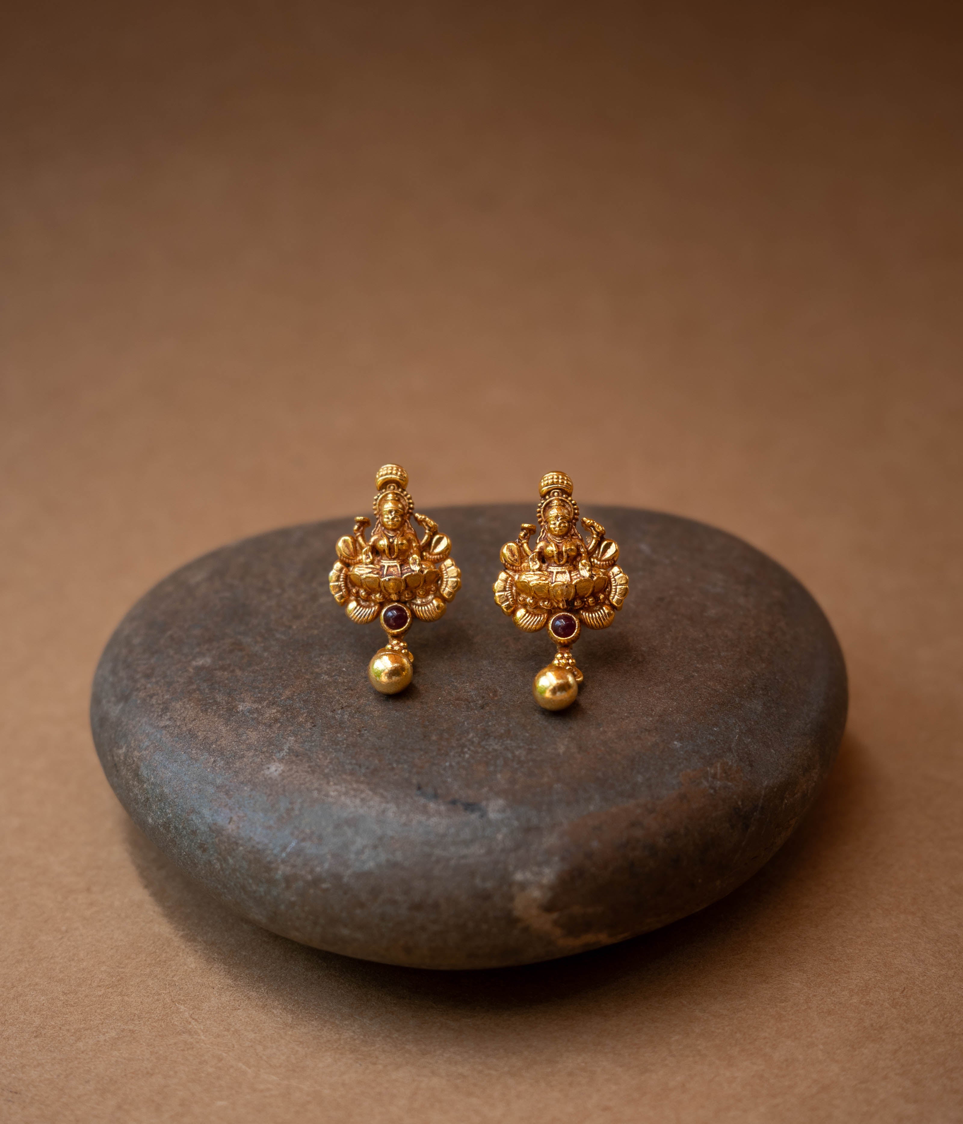 Traditional stud earrings with a goddess figure, a red stone, and a dangling bead.
