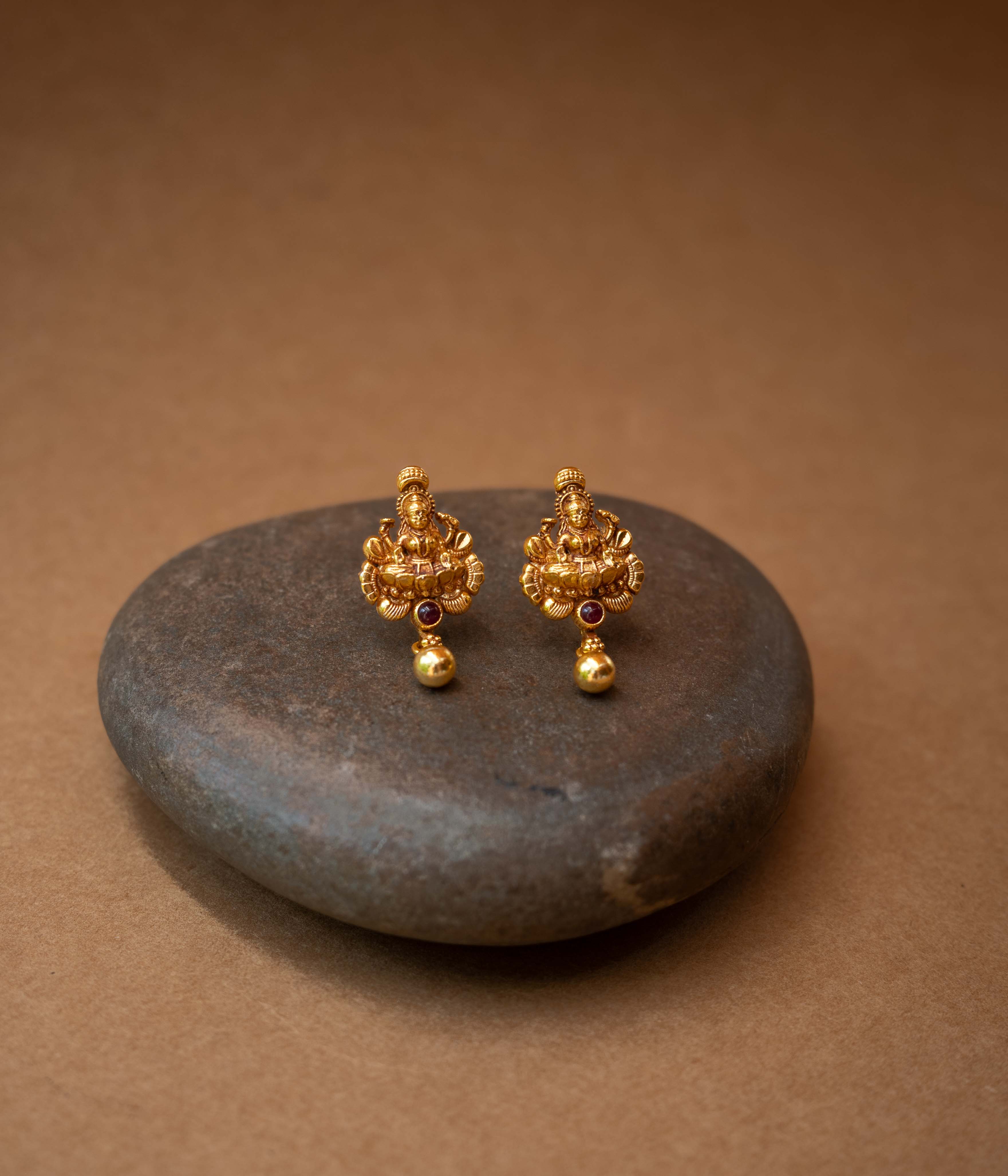 Traditional stud earrings with a goddess figure, a red stone, and a dangling bead.