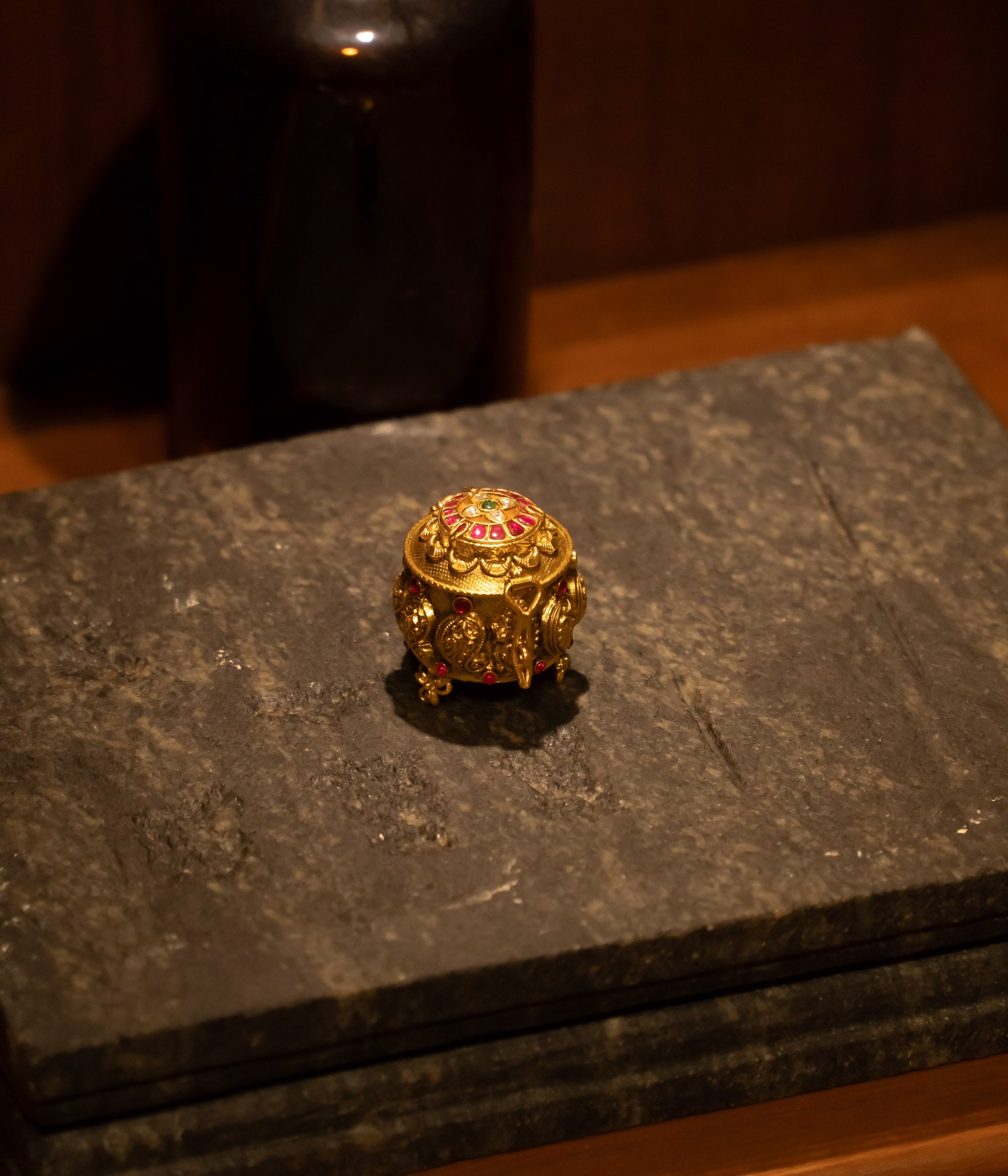 Temple-style golden kumkum box with red and green Kundan stones, placed on stone surface.