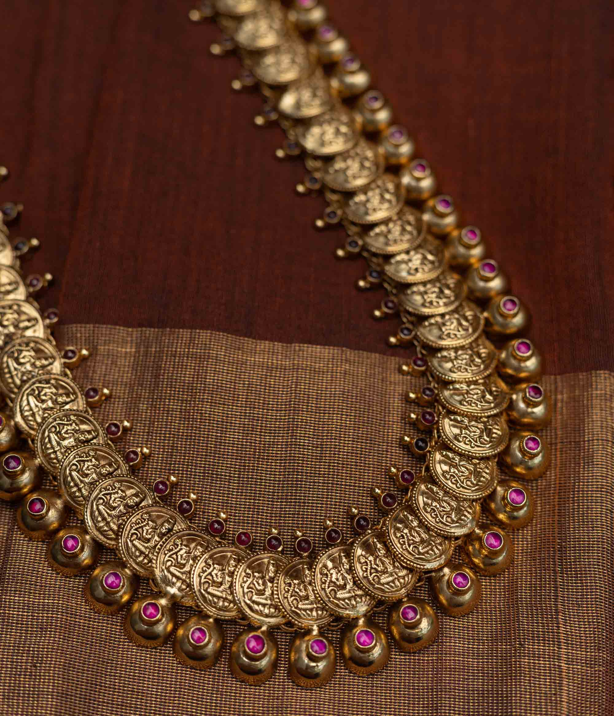 Long Kasu Mala necklace featuring an array of antique-finished gold (likely 92.5 sterling silver) coins depicting Goddess Lakshmi, delicately accented with ruby-toned stones, a timeless symbol of blessings and cultural richness.
