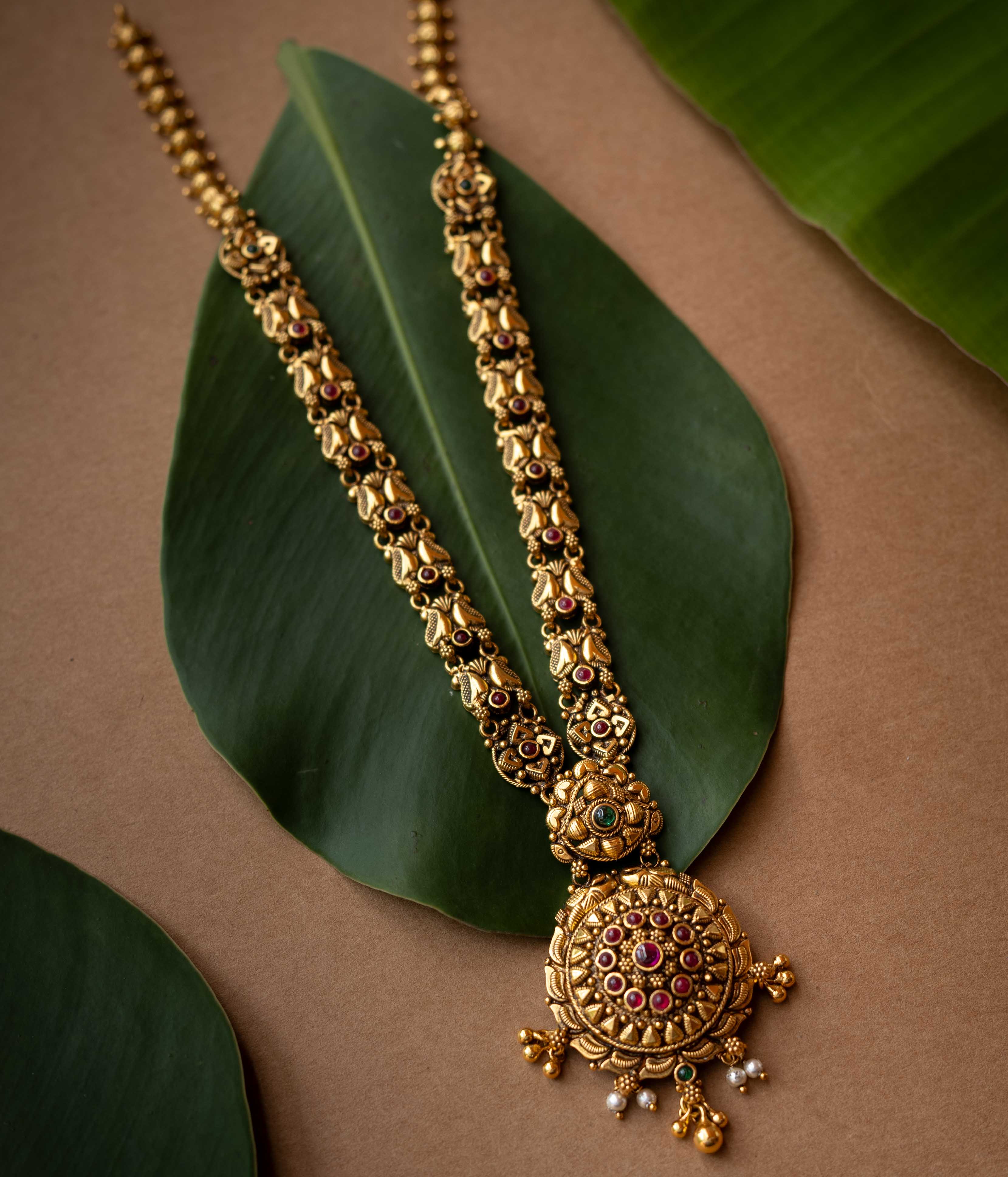 Antique gold temple necklace with lotus motifs and ruby-green stone accents from Temple Collection.

