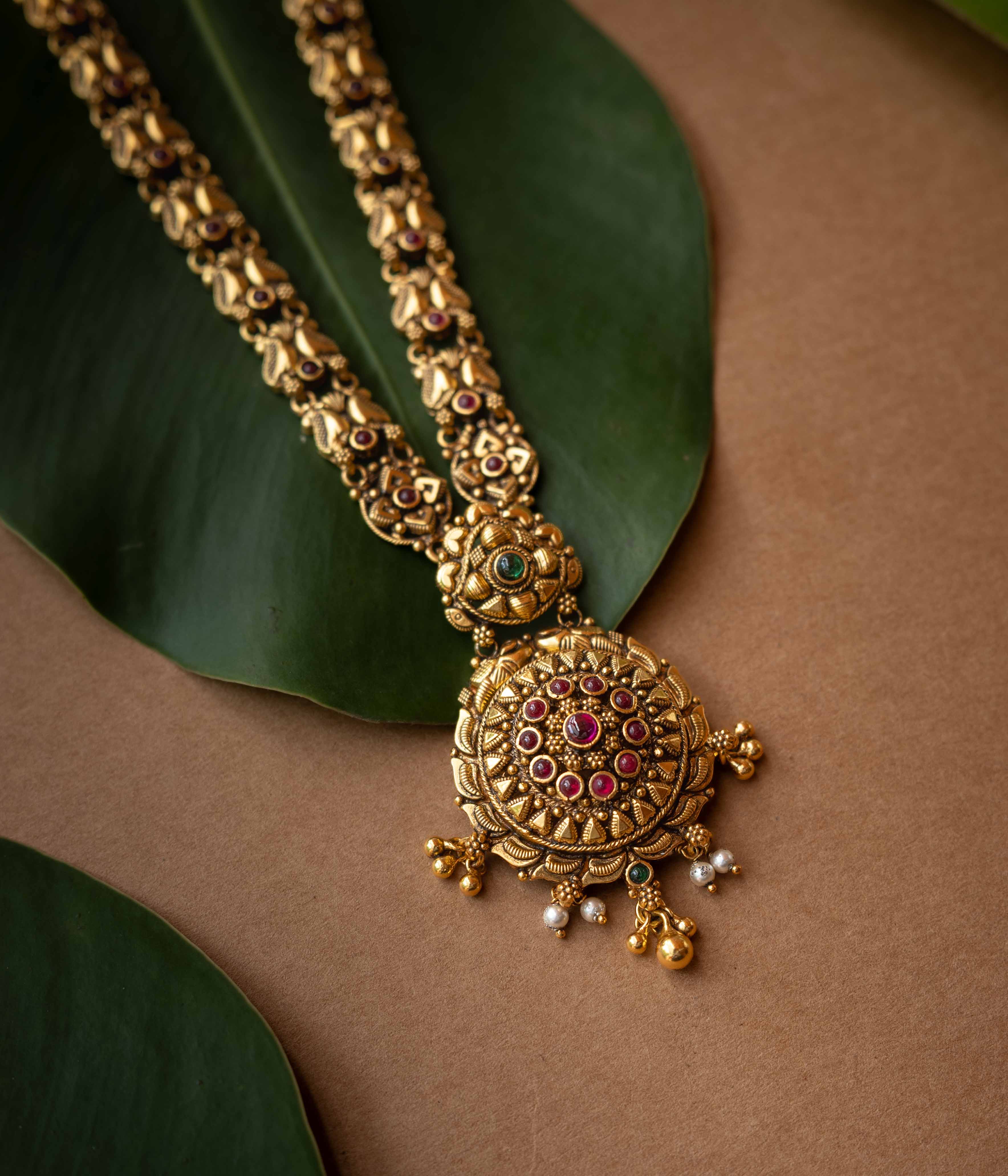 Antique gold temple necklace with lotus motifs and ruby-green stone accents from Temple Collection.
