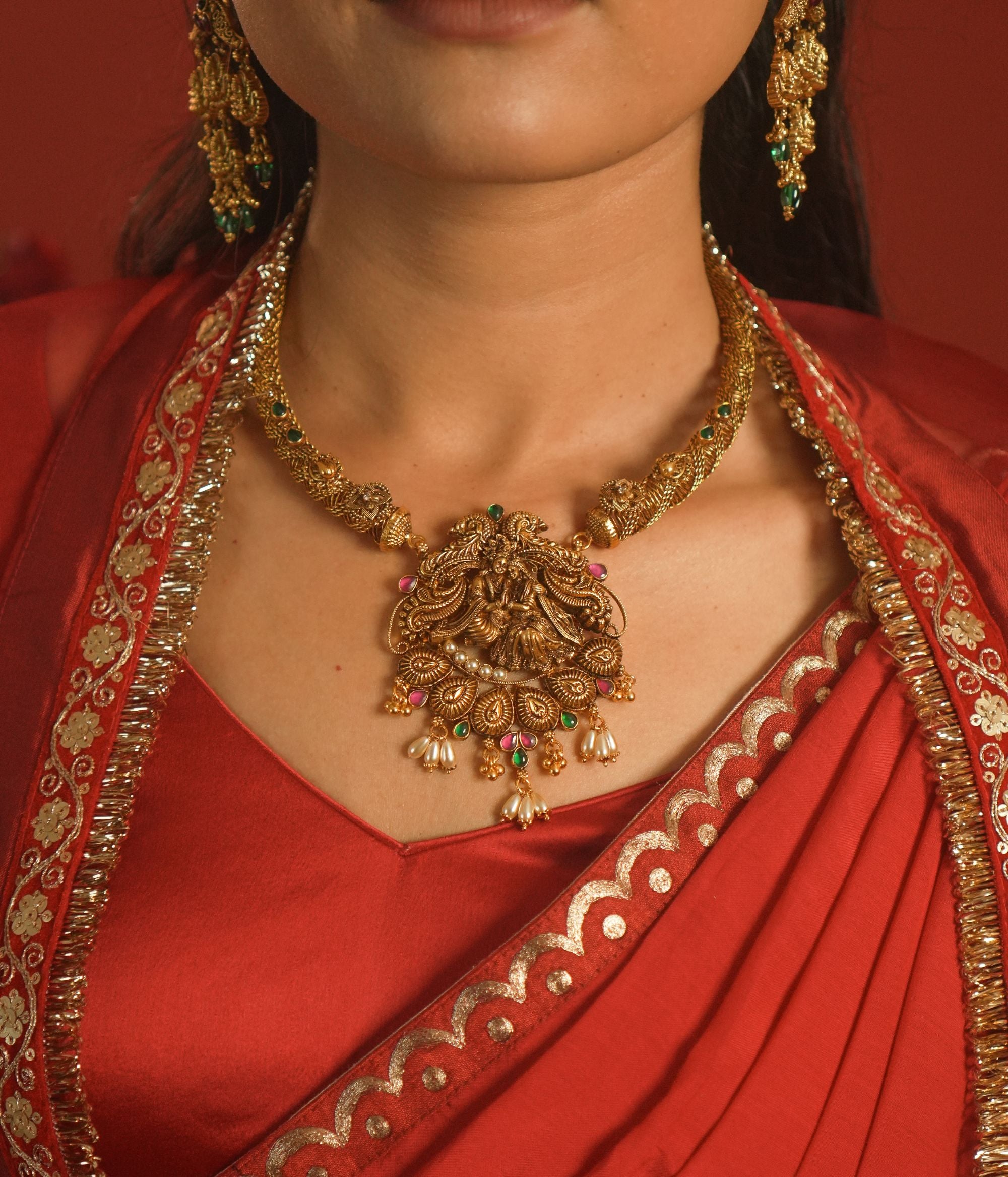 Woman wearing a red saree with gold jewelry, including a necklace and earrings.