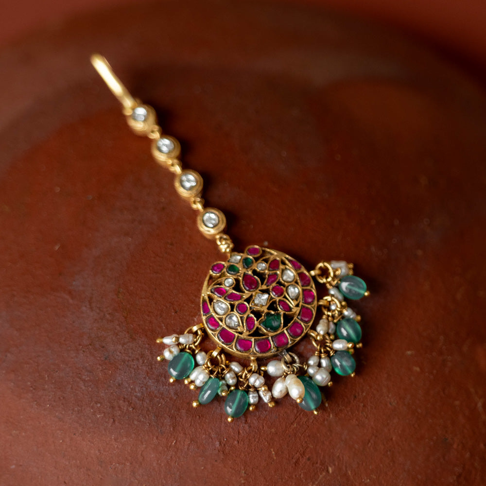 A handcrafted 92.5 sterling silver and 22ct gold-plated Kundan tikka, featuring red, green, and white Kundan stones, green beads, and pearls, showcasing intricate gold detailing and evoking timeless regal charm.