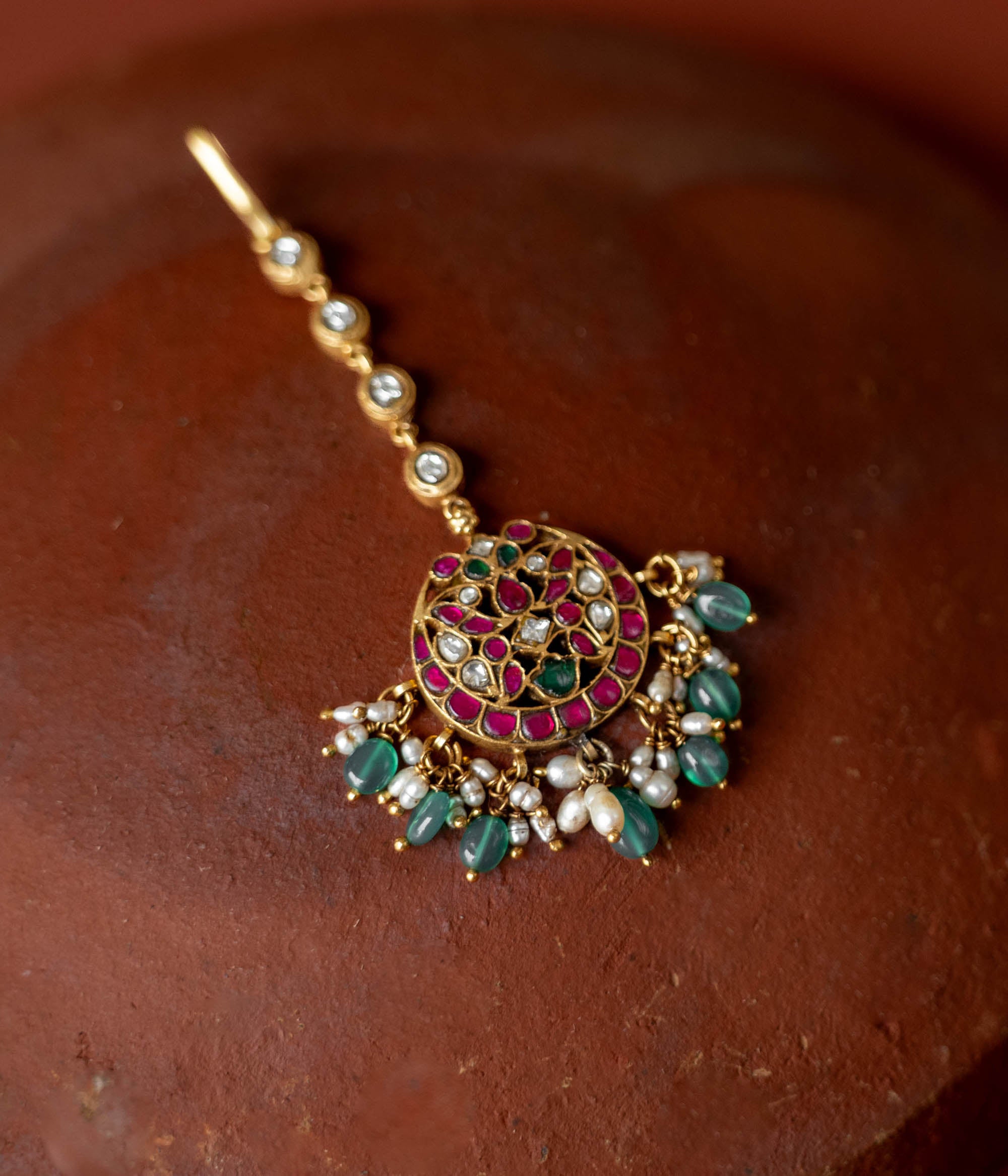 A handcrafted 92.5 sterling silver and 22ct gold-plated Kundan tikka, featuring red, green, and white Kundan stones, green beads, and pearls, showcasing intricate gold detailing and evoking timeless regal charm.