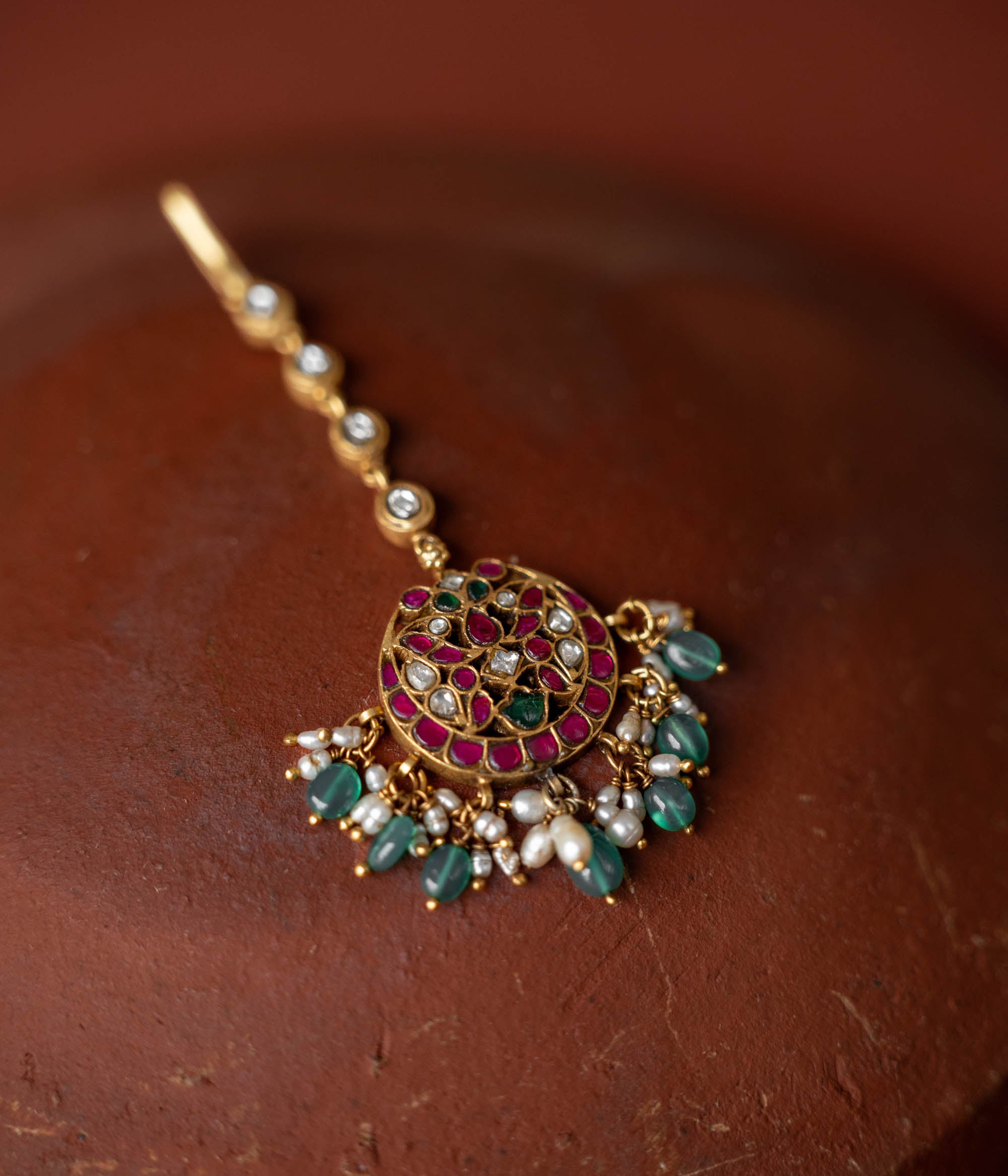 A handcrafted 92.5 sterling silver and 22ct gold-plated Kundan tikka, featuring red, green, and white Kundan stones, green beads, and pearls, showcasing intricate gold detailing and evoking timeless regal charm.