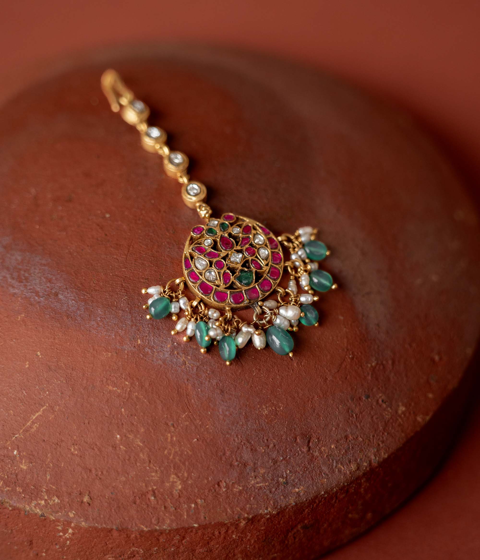 A handcrafted 92.5 sterling silver and 22ct gold-plated Kundan tikka, featuring red, green, and white Kundan stones, green beads, and pearls, showcasing intricate gold detailing and evoking timeless regal charm.