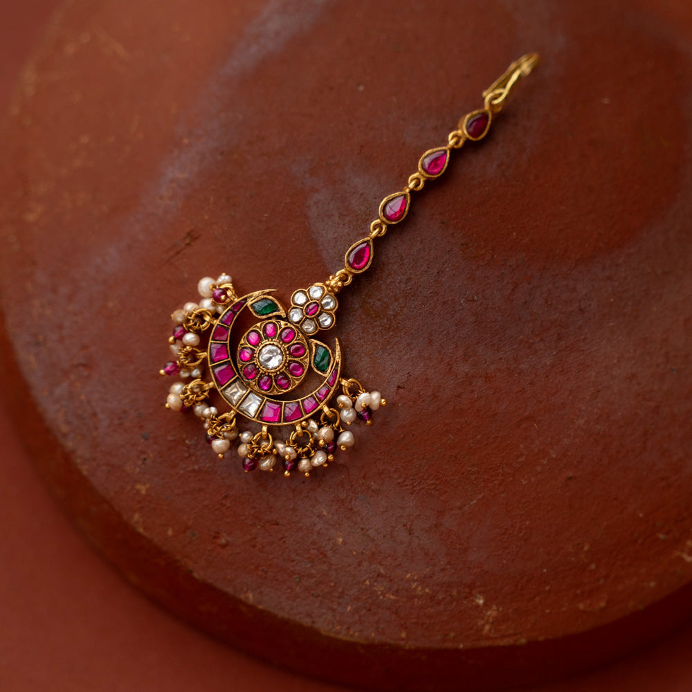 A handcrafted 92.5 sterling silver and 22ct gold-plated Kundan tikka, featuring red, green, and white Kundan stones and pearls, with intricate gold detailing, inspired by the glow of the moon and radiating celestial grace and timeless charm.