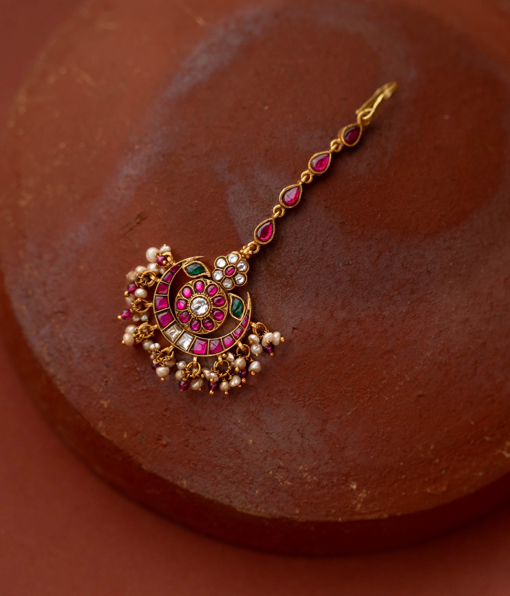 A handcrafted 92.5 sterling silver and 22ct gold-plated Kundan tikka, featuring red, green, and white Kundan stones and pearls, with intricate gold detailing, inspired by the glow of the moon and radiating celestial grace and timeless charm.