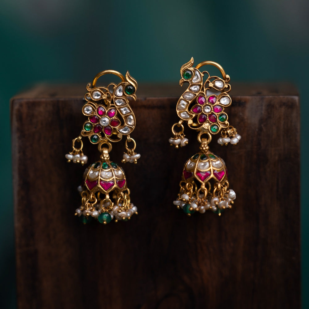 A pair of handcrafted 92.5 sterling silver earrings with screw fastenings, featuring a regal peacock motif adorned with red, white, and green Kundan stones, green beads, and pearls, showcasing intricate gold detailing and capturing the essence of grace and tradition.