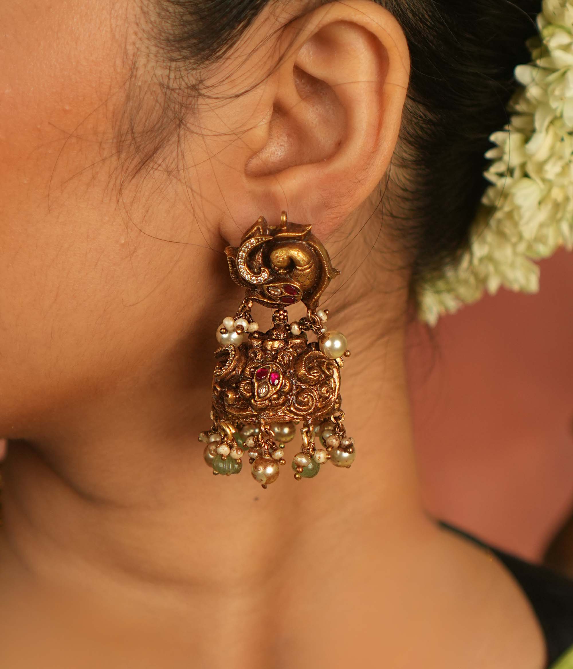 Temple whispers reborn! These antique gold jhumka earrings feature intricate floral designs adorned with radiant red and white Kundan stones. Graceful green and white pearl drops enhance the charm, echoing divine artistry and timeless cultural heritage.