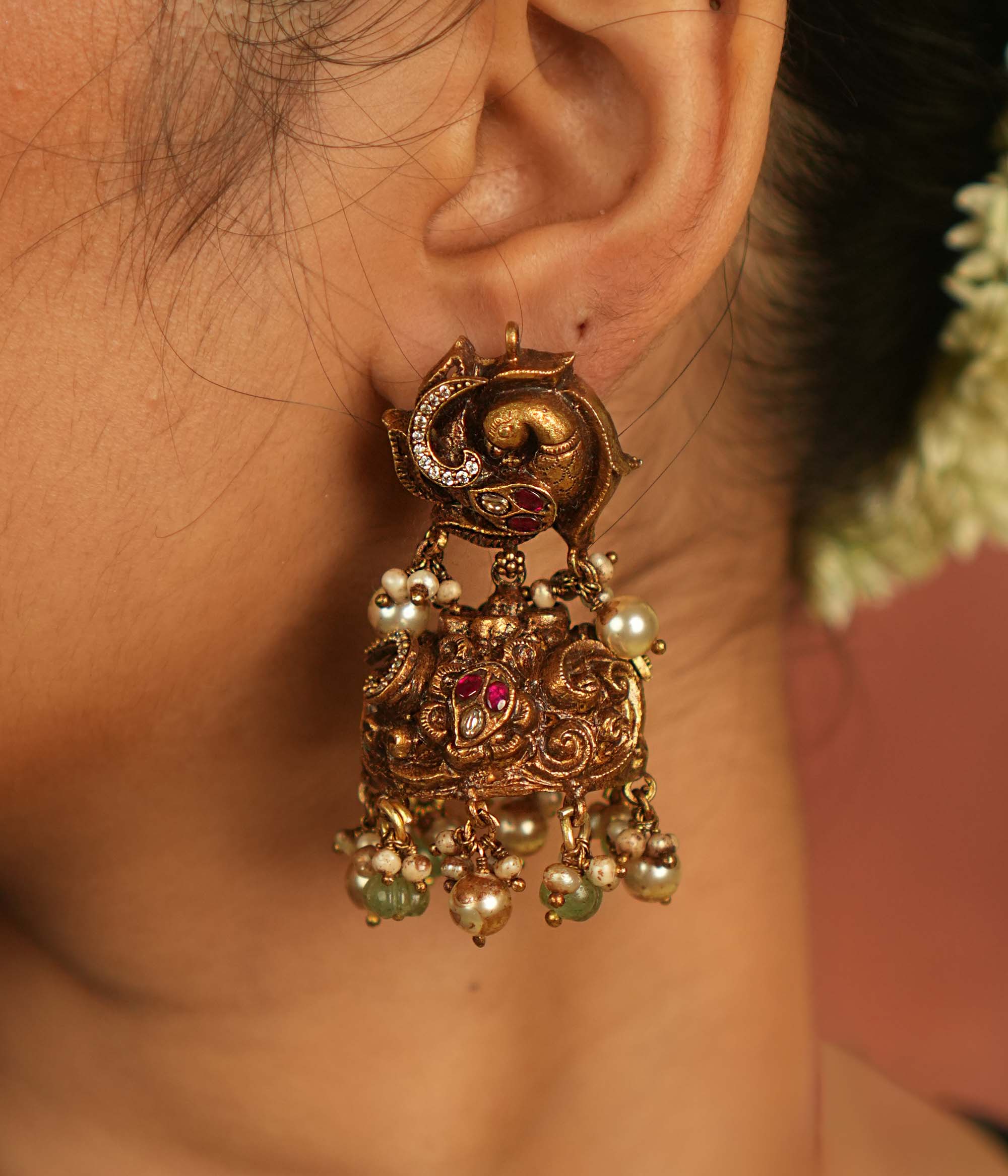 Temple whispers reborn! These antique gold jhumka earrings feature intricate floral designs adorned with radiant red and white Kundan stones. Graceful green and white pearl drops enhance the charm, echoing divine artistry and timeless cultural heritage.