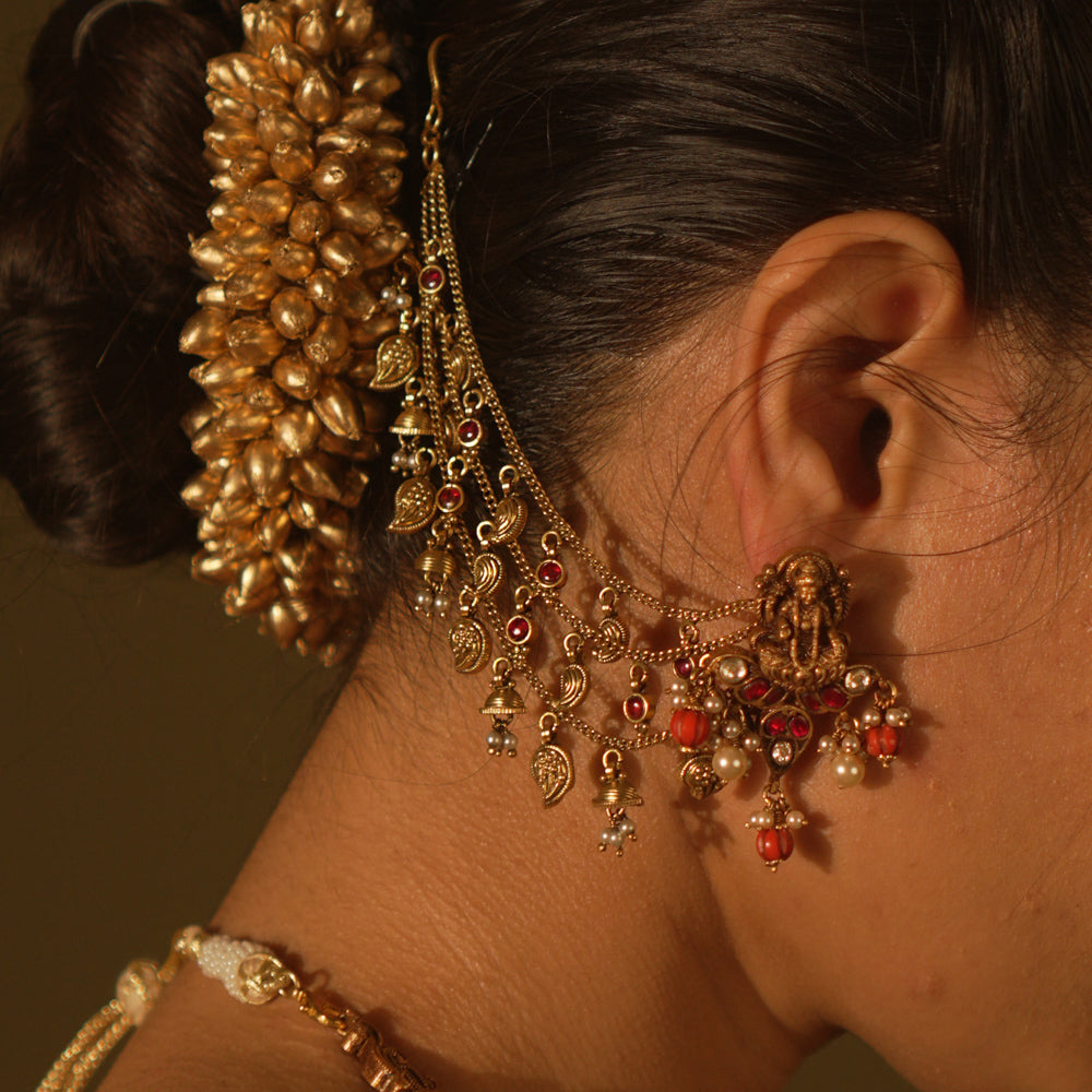 Antique gold temple earrings with a goddess figure, red and green kundan, red beads, and pearls, with multi-strand chains extending to the hair.