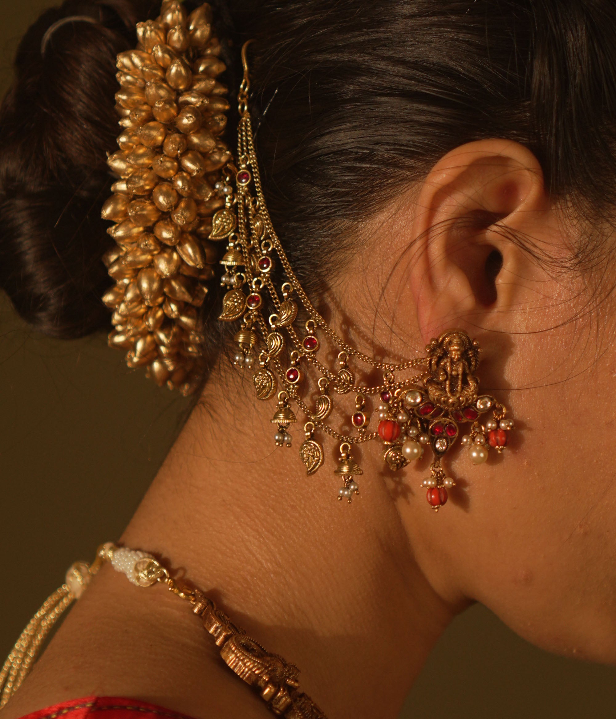 Antique gold temple earrings with a goddess figure, red and green kundan, red beads, and pearls, with multi-strand chains extending to the hair.