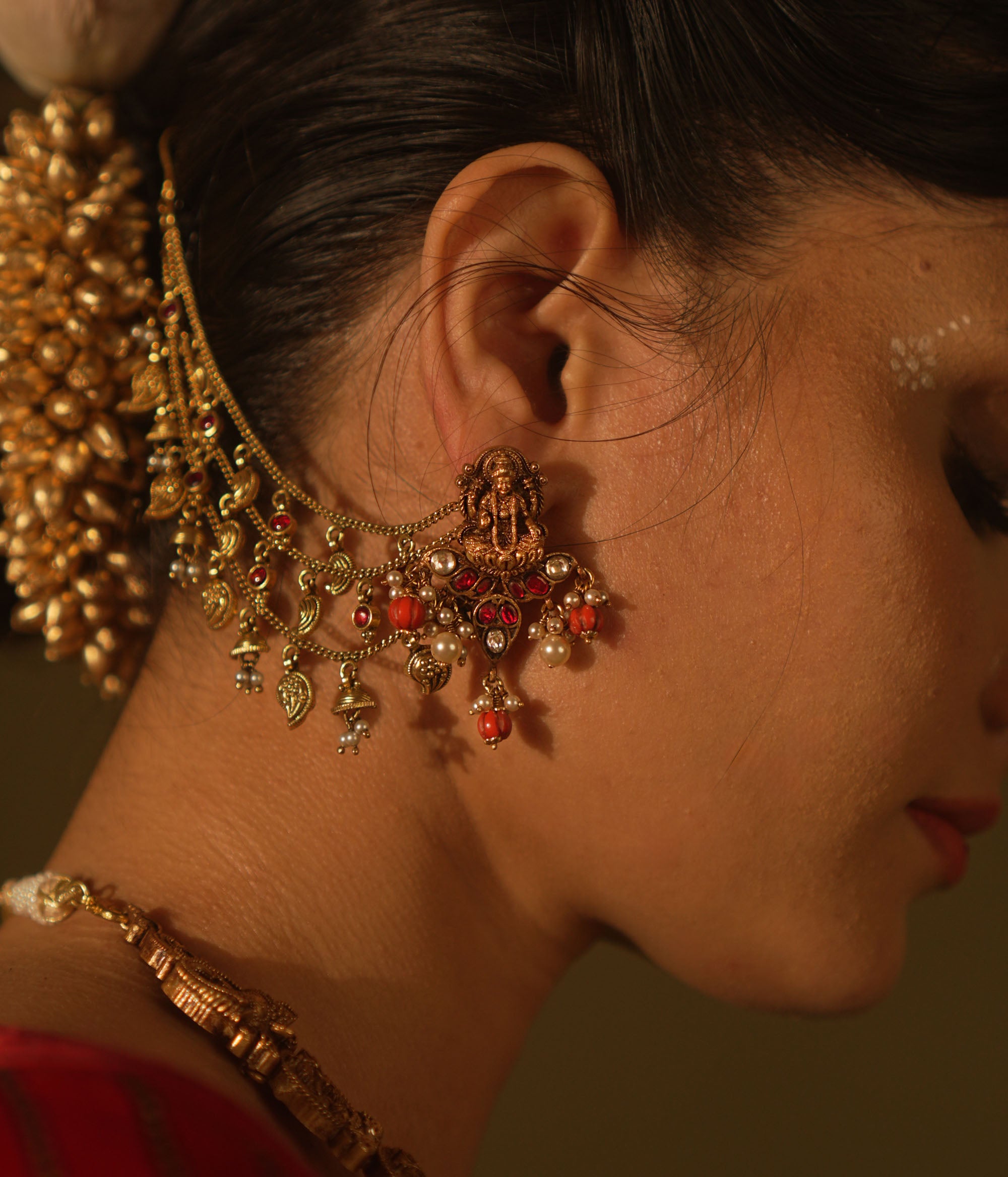 Antique gold temple earrings with a goddess figure, red and green kundan, red beads, and pearls, with multi-strand chains extending to the hair.