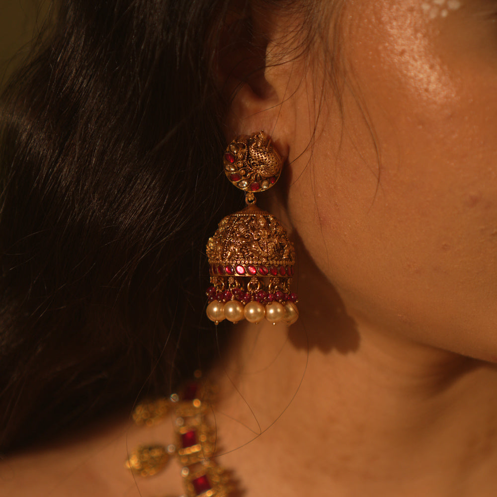 Antique gold temple jhumka earrings with peacock motif, red and green kundan, red beads, and pearl drops, on a woman's ear.