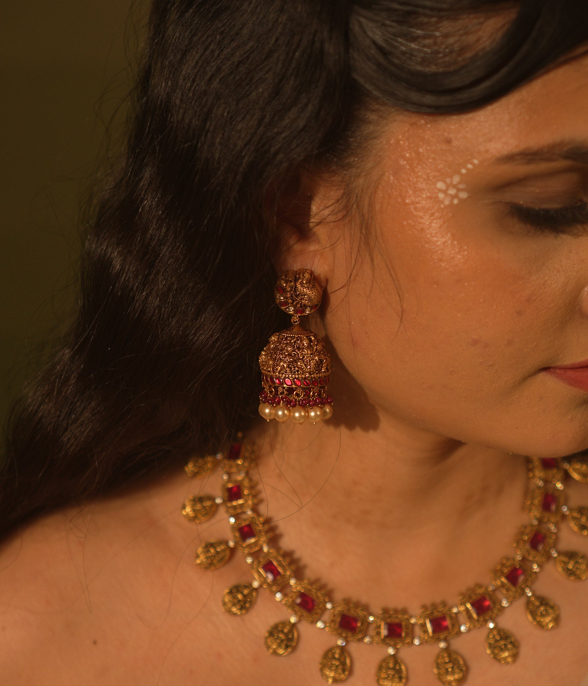 Antique gold temple jhumka earrings with peacock motif, red and green kundan, red beads, and pearl drops, on a woman's ear.
