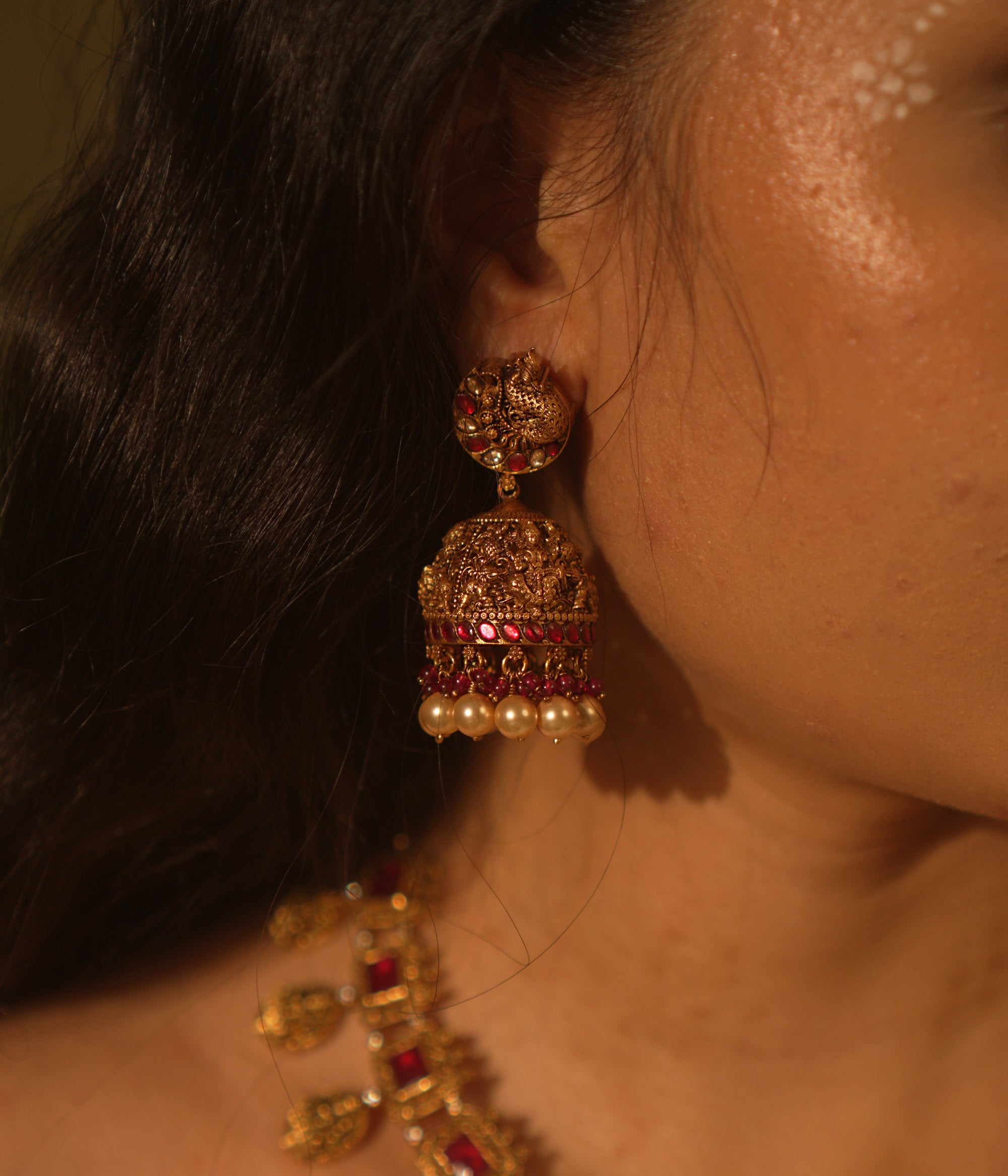 Antique gold temple jhumka earrings with peacock motif, red and green kundan, red beads, and pearl drops, on a woman's ear.