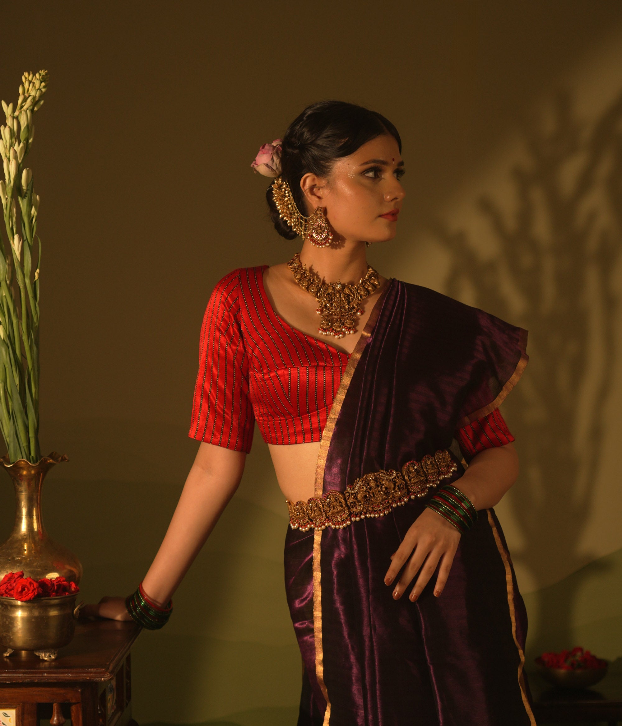 Temple vaddanam with goddess motifs, red-white Kundan stones, and pearl drops on a saree.