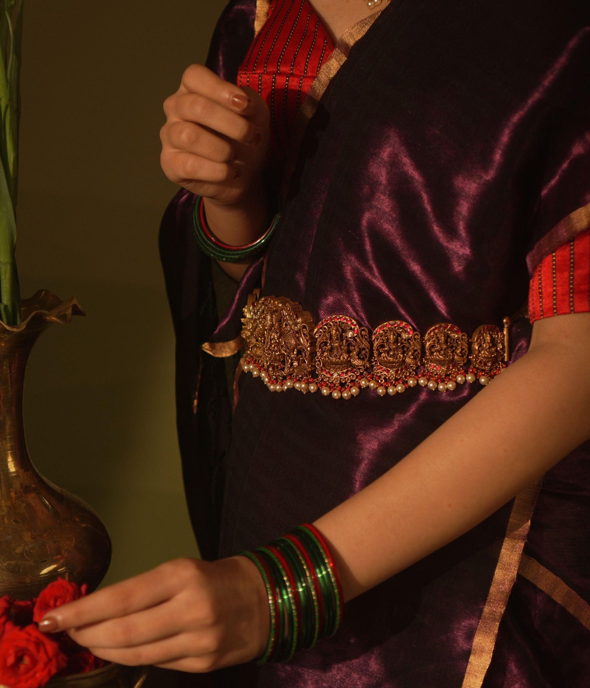Temple vaddanam with goddess motifs, red-white Kundan stones, and pearl drops on a saree.
