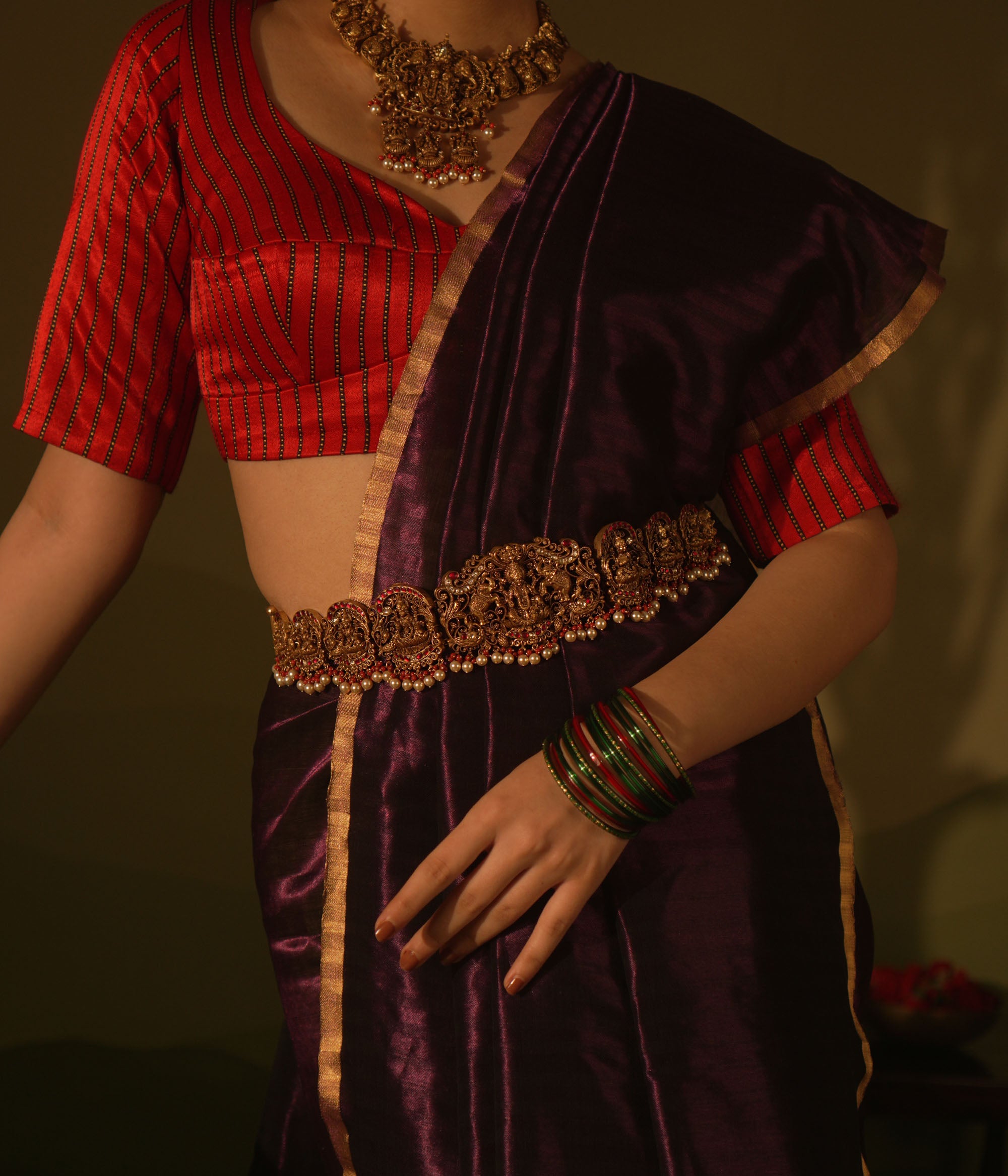 Temple vaddanam with goddess motifs, red-white Kundan stones, and pearl drops on a saree.