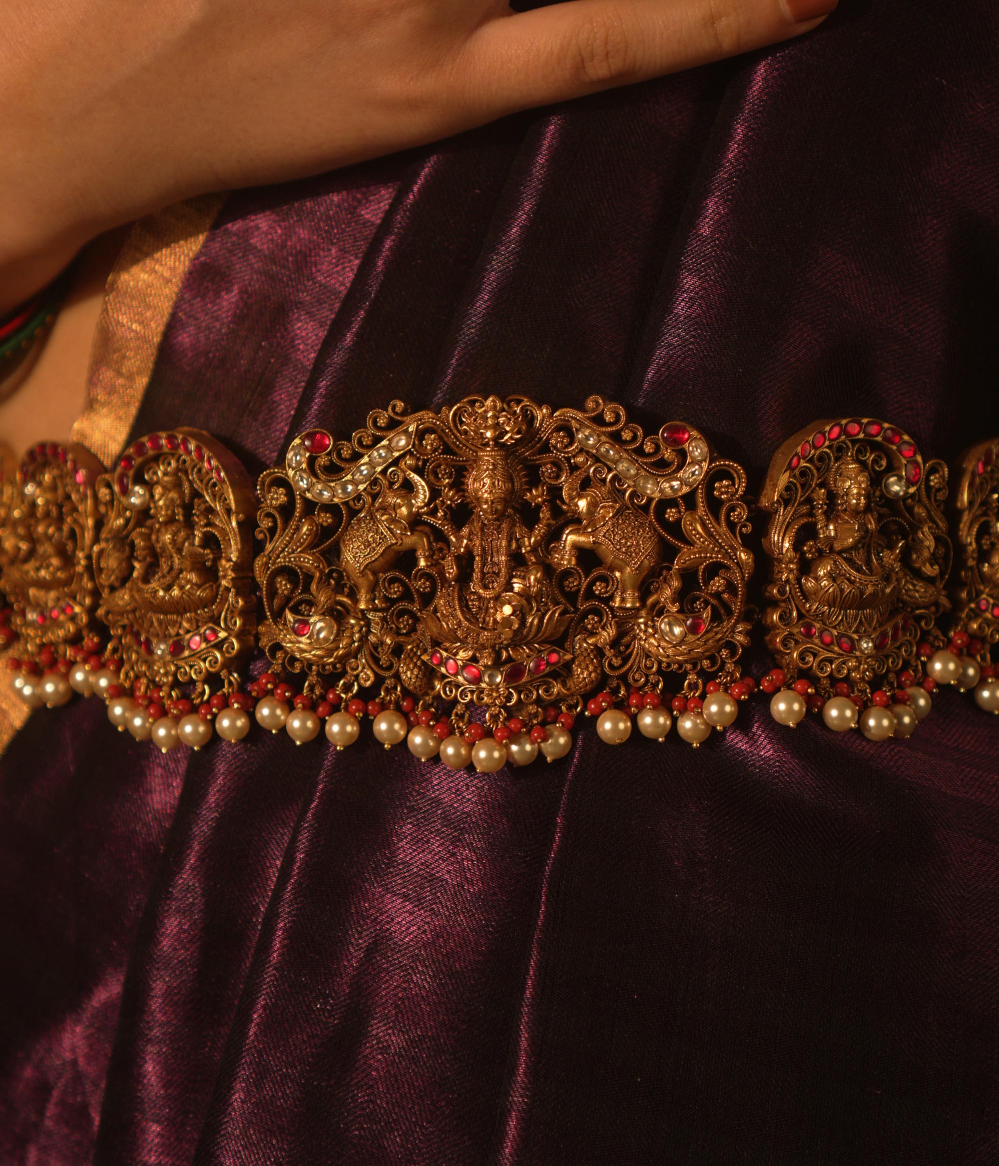 Temple vaddanam with goddess motifs, red-white Kundan stones, and pearl drops on a saree.