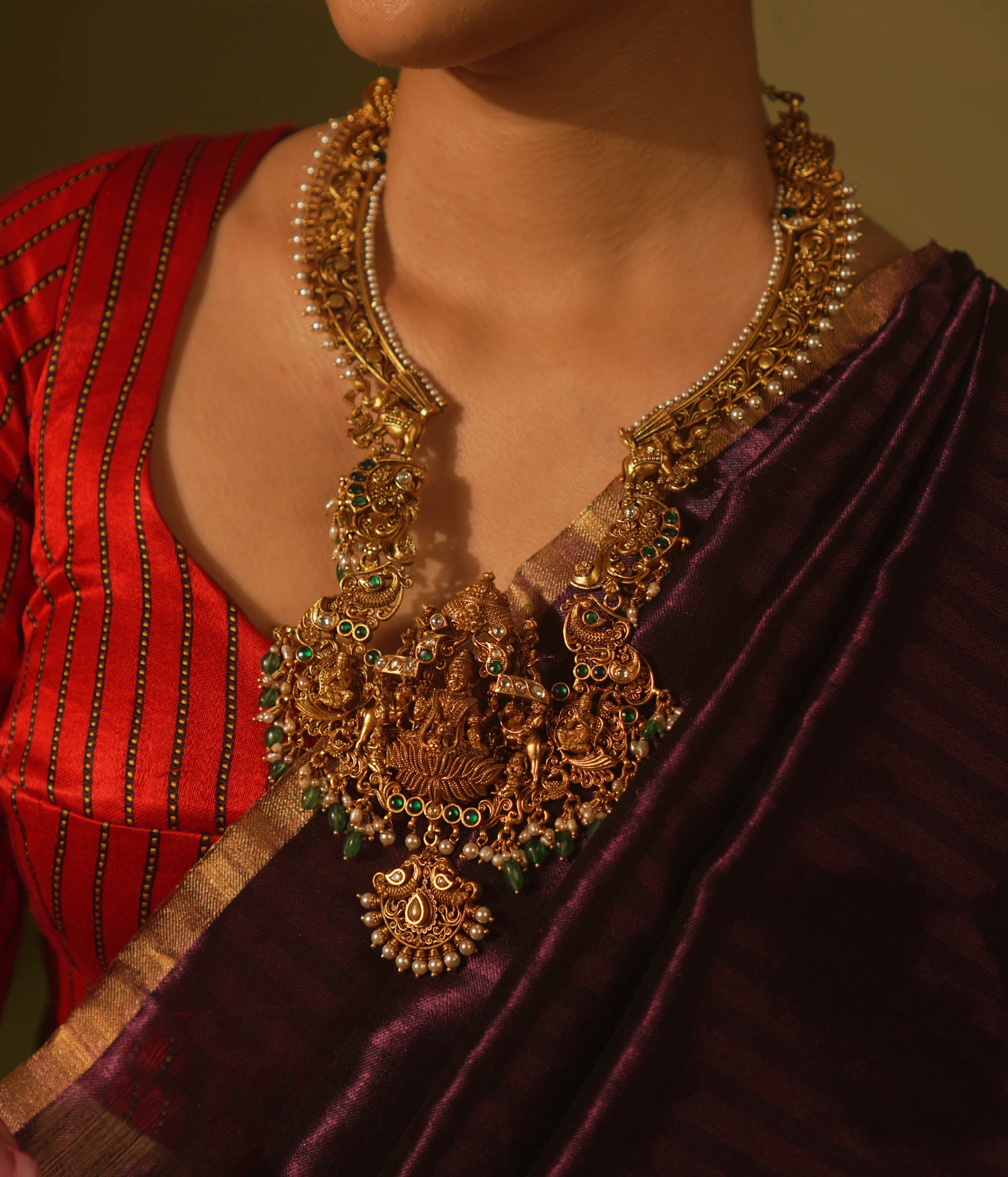 Temple-style gold necklace featuring Goddess Lakshmi, green beads, white Kundan stones, and intricate divine motifs in antique finish.