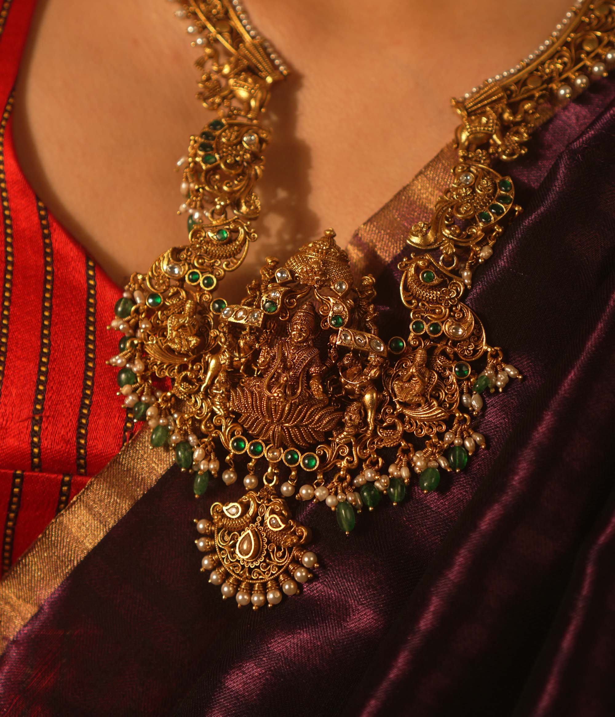 Temple-style gold necklace featuring Goddess Lakshmi, green beads, white Kundan stones, and intricate divine motifs in antique finish.