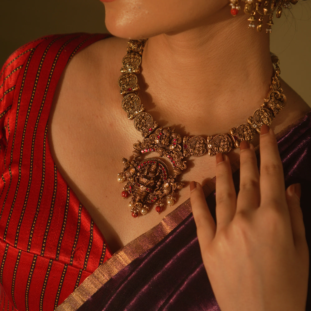 Antique gold temple necklace with goddess motifs, red and white kundan, red beads, and pearl drops, worn by a woman.