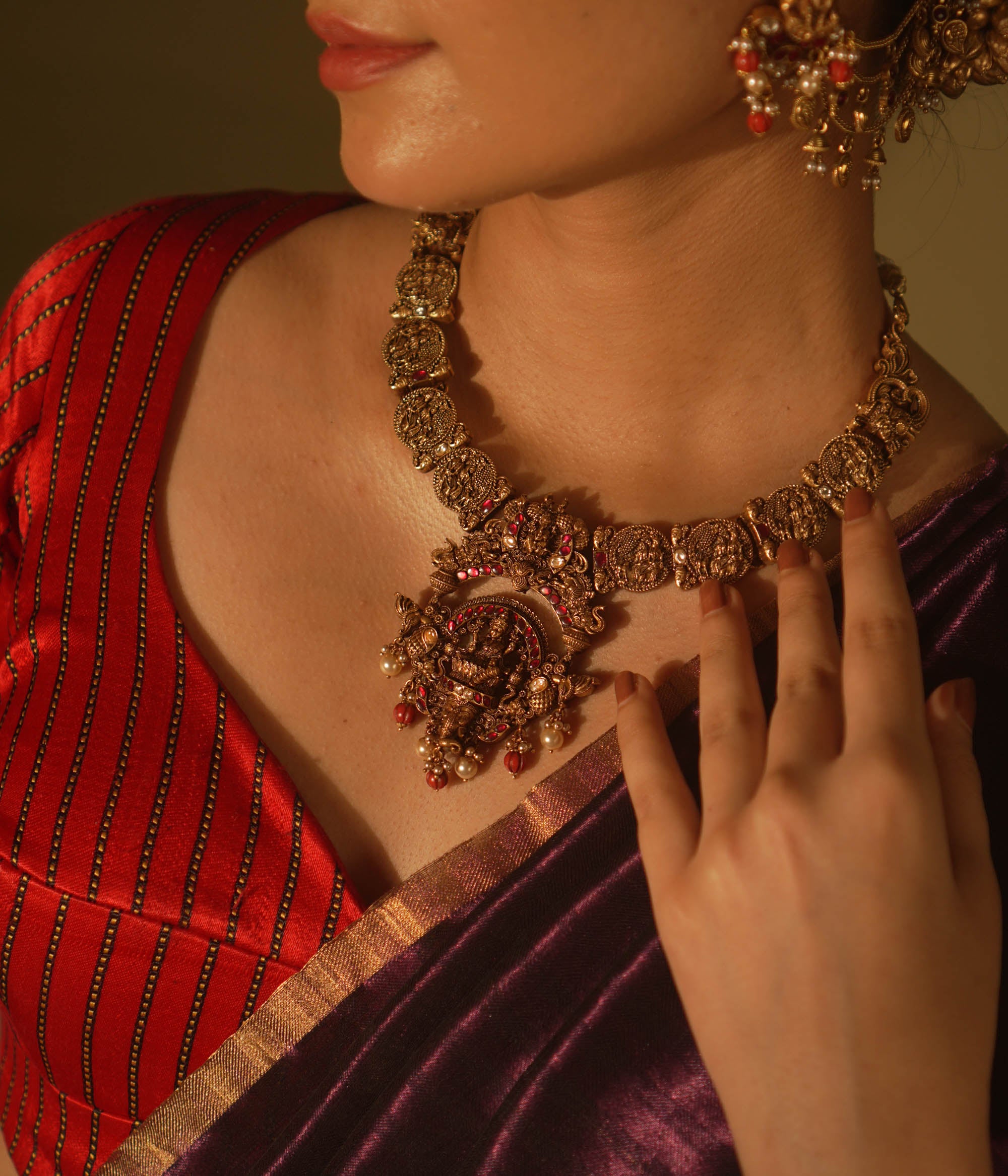 Antique gold temple necklace with goddess motifs, red and white kundan, red beads, and pearl drops, worn by a woman.