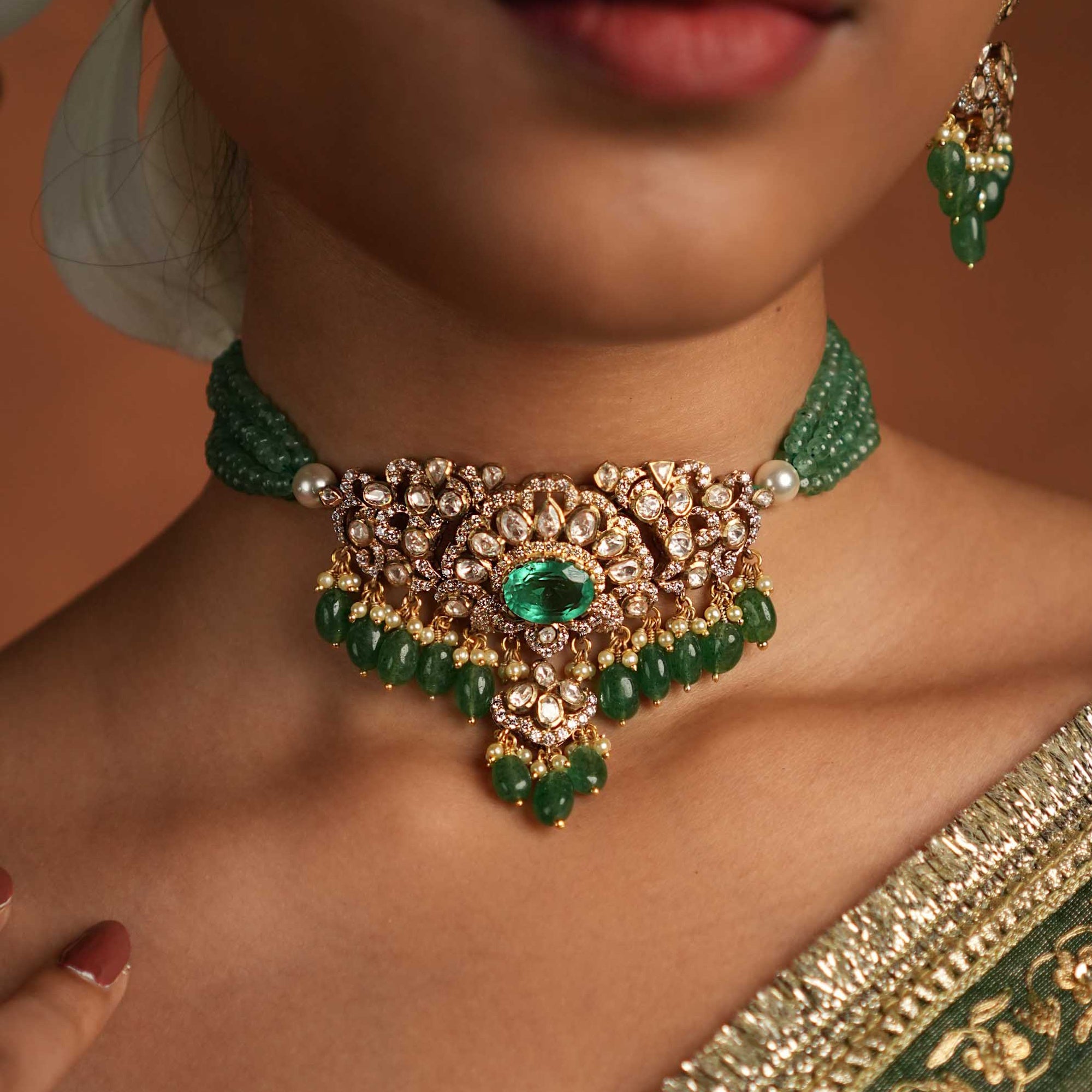 Close-up of a model wearing an ornate gold-toned choker with a central green stone, emerald bead strands, pearl accents, and dangling green bead drops, styled with a traditional outfit.