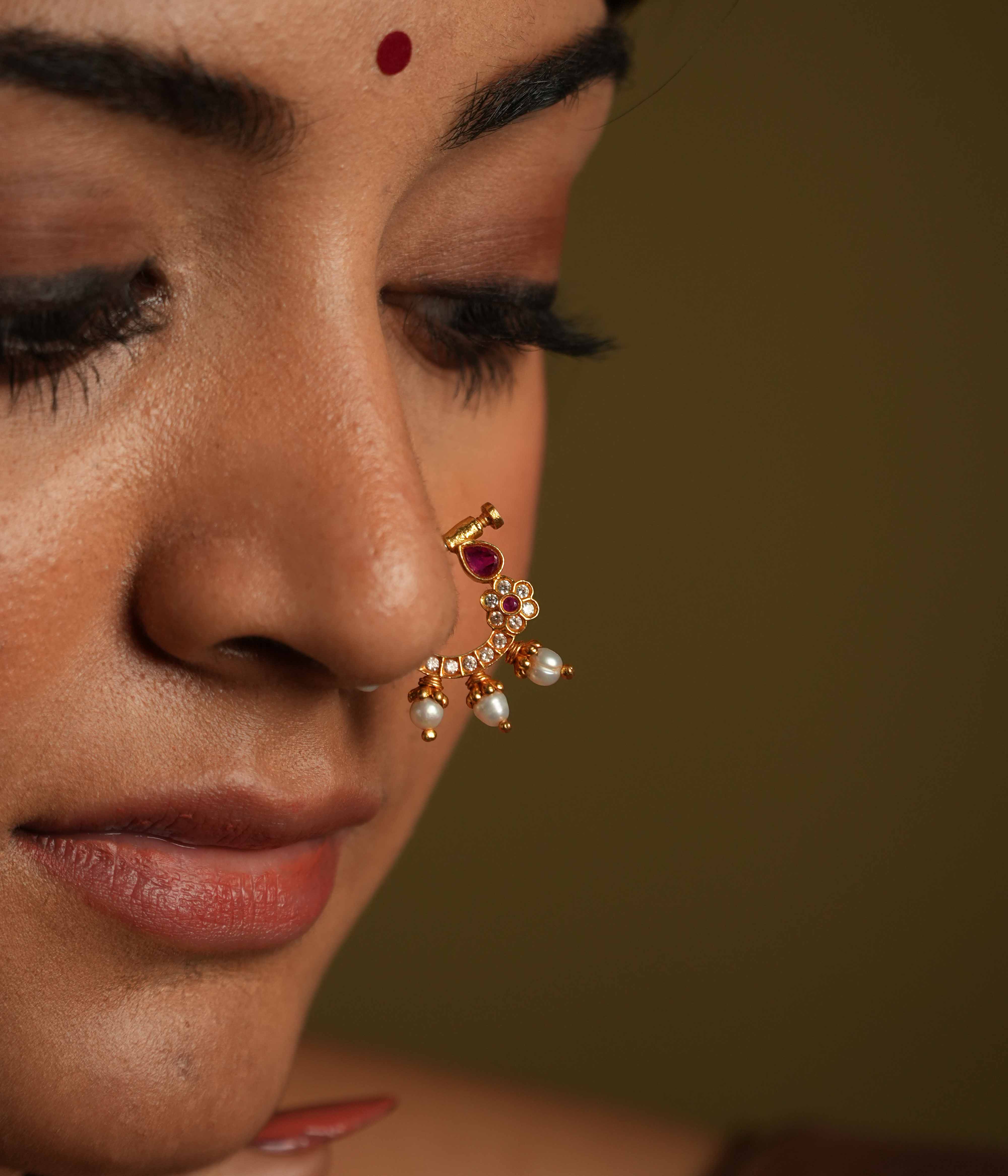 A gold-plated 92.5 sterling silver nose ring featuring a vintage South Indian-inspired design. It is adorned with CZ stones and red cut stones, set in a white Kundan setting. The piece includes a minimal flower motif with three bold pearl edgings, exuding timeless elegance and classic sophistication.