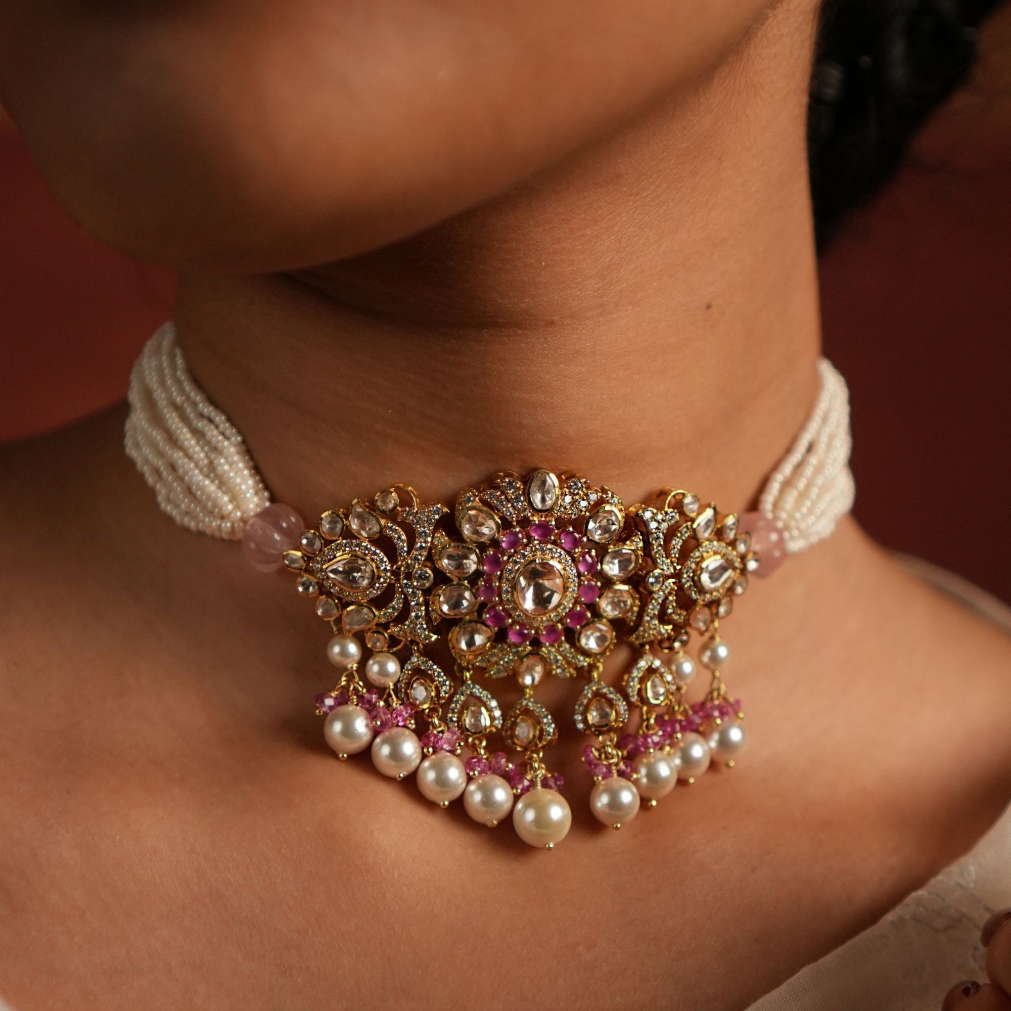 Close-up of a model wearing a gold-toned choker with pink stone detailing, pearl bead strands, and dangling pearl drops against a traditional outfit.